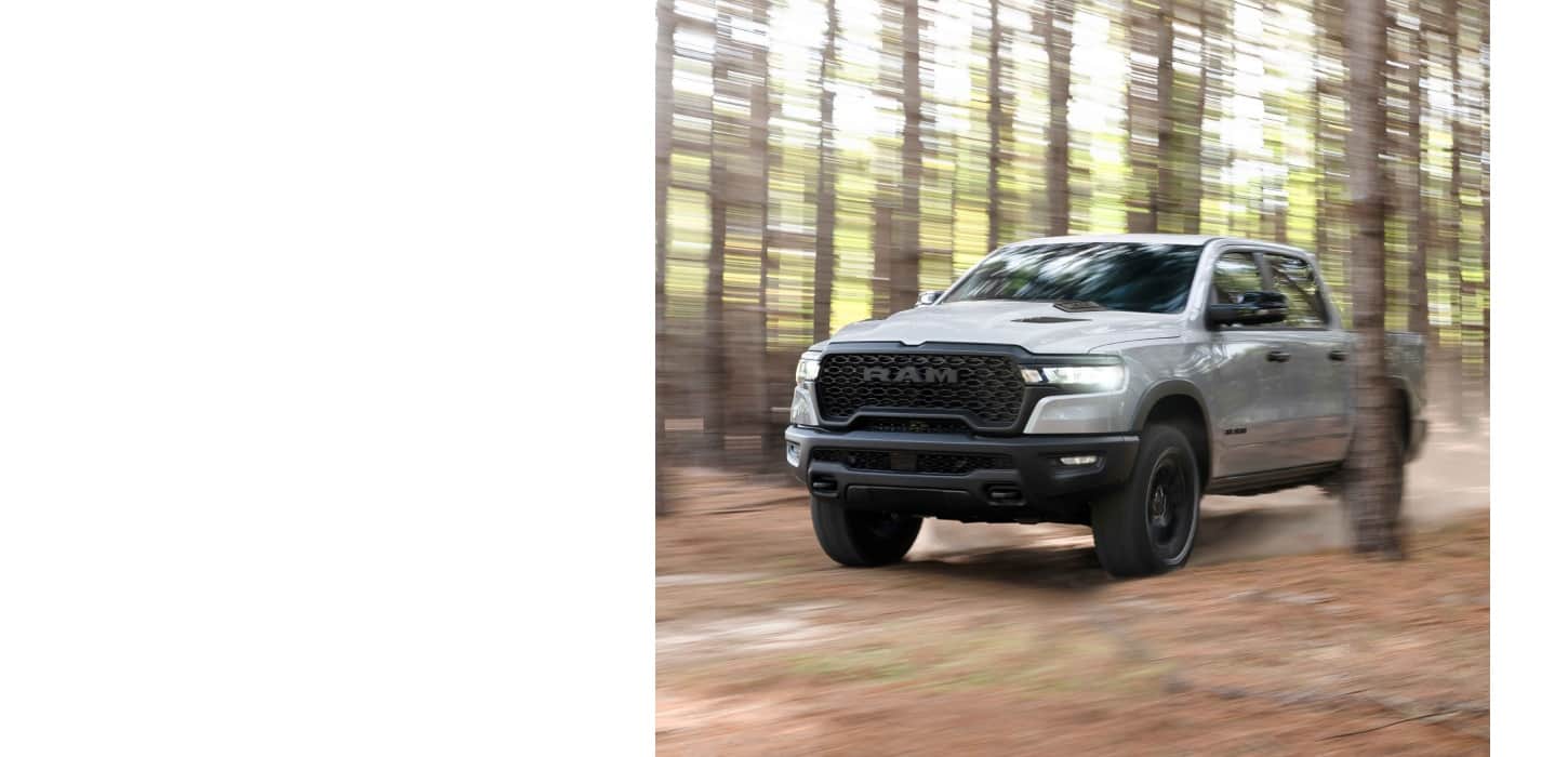A white 2026 Ram 1500 Rebel 4x4 Crew Cab traveling through a forest with the background blurred.