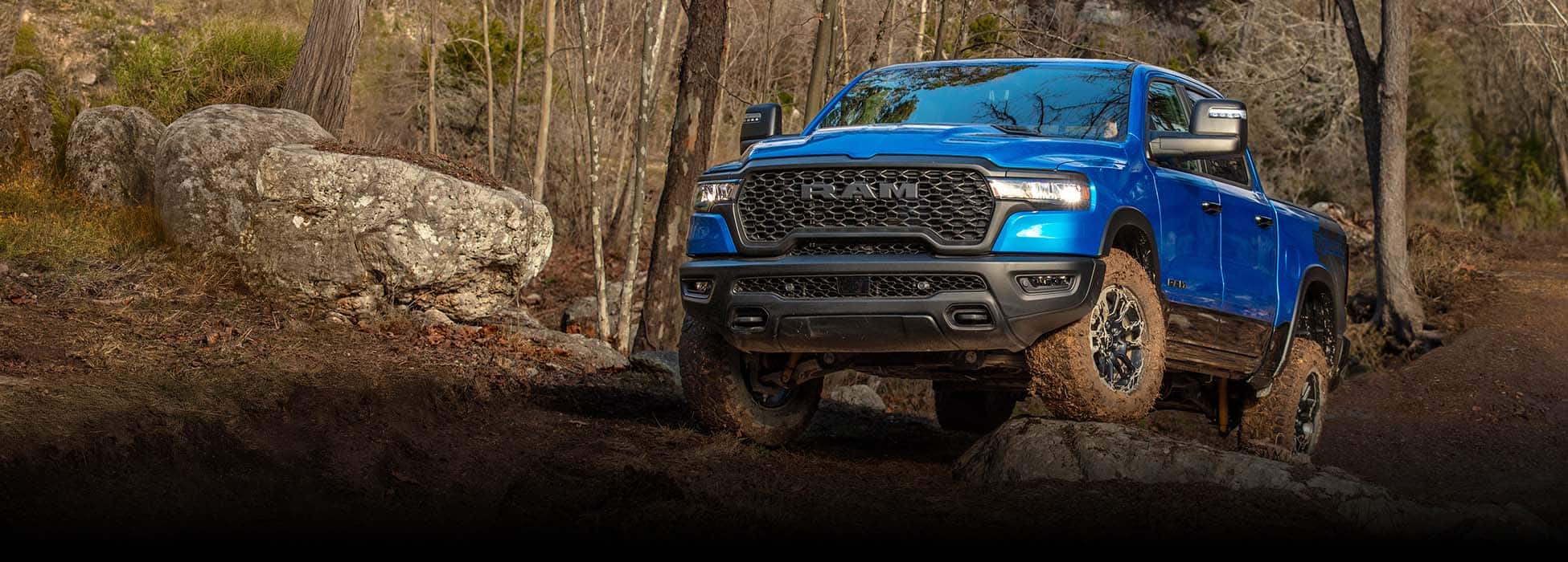 A blue 2026 Ram 1500 Rebel 4x4 Crew Cab climbing a rocky trail off-road, with a forest in the background.