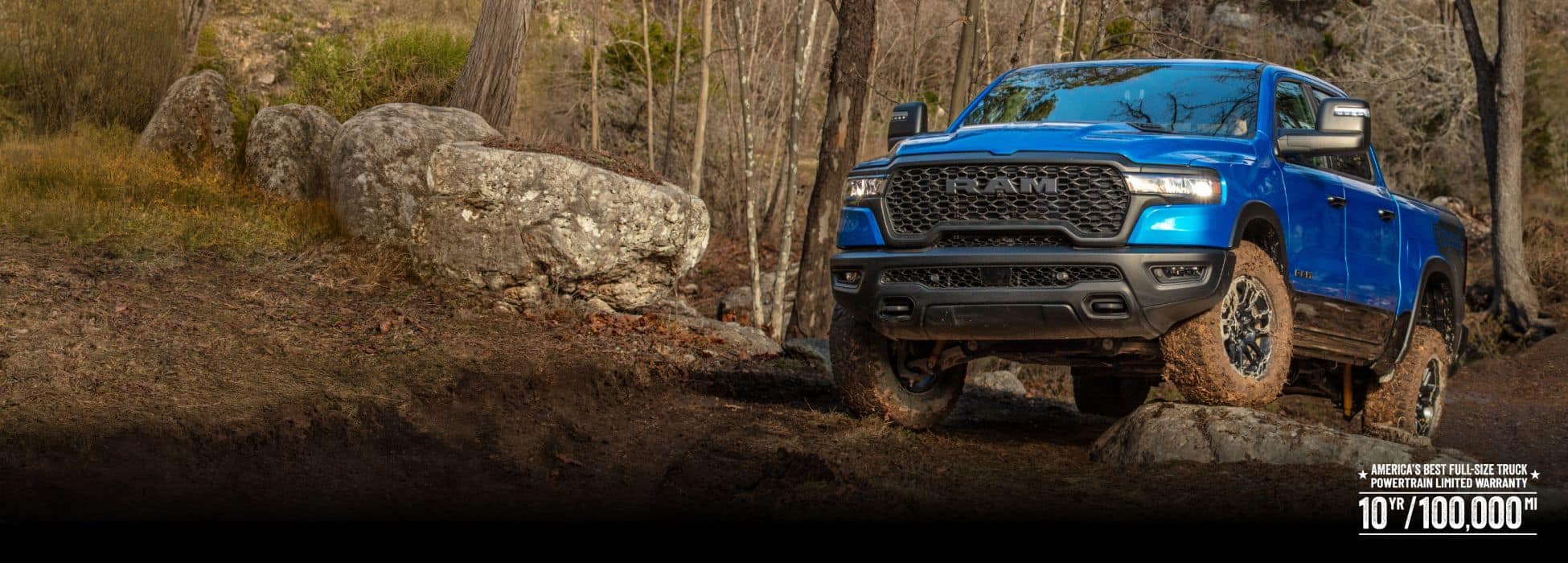 A blue 2026 Ram 1500 Rebel 4x4 Crew Cab climbing a rocky trail off-road, with a forest in the background.