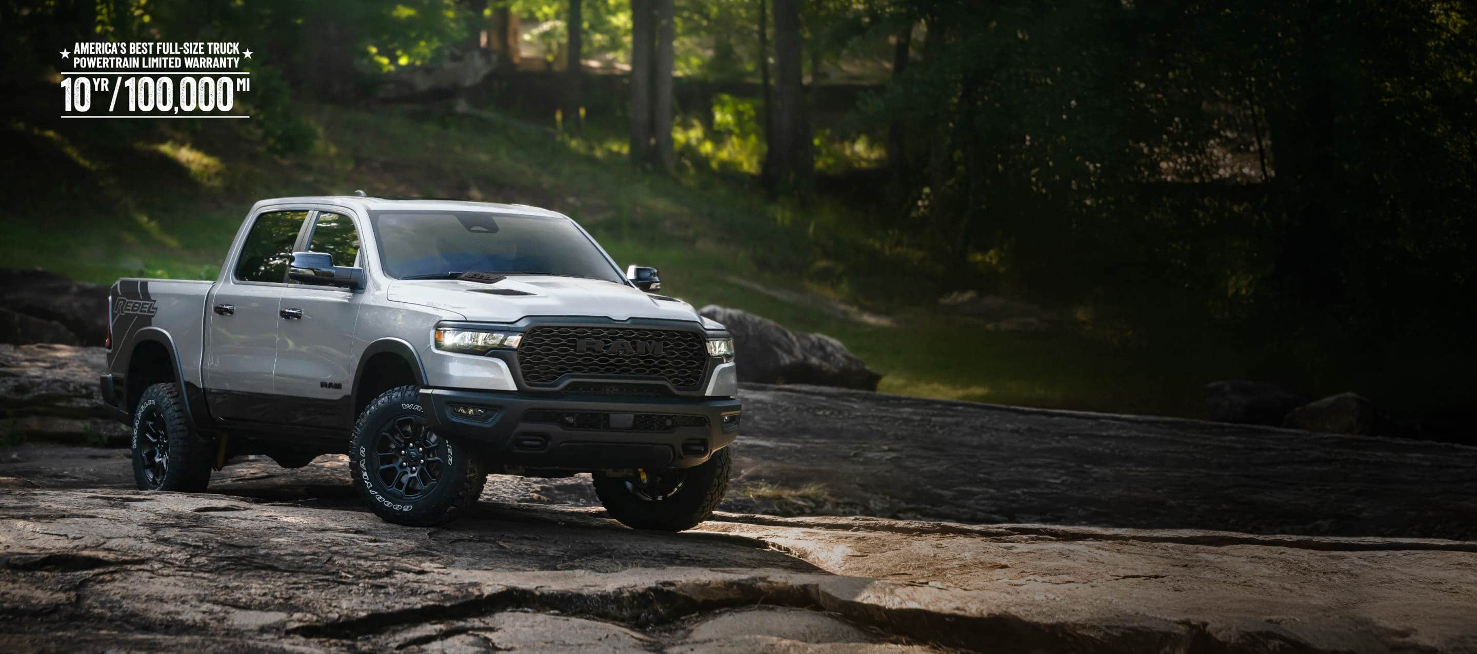 A white 2026 Ram 1500 Rebel 4x4 Crew Cab traveling down a rocky trail off-road, with a forest in the background. America's Best Full-Size Truck Powertrain Limited Warranty. 10-Year, 100,000-Mile.