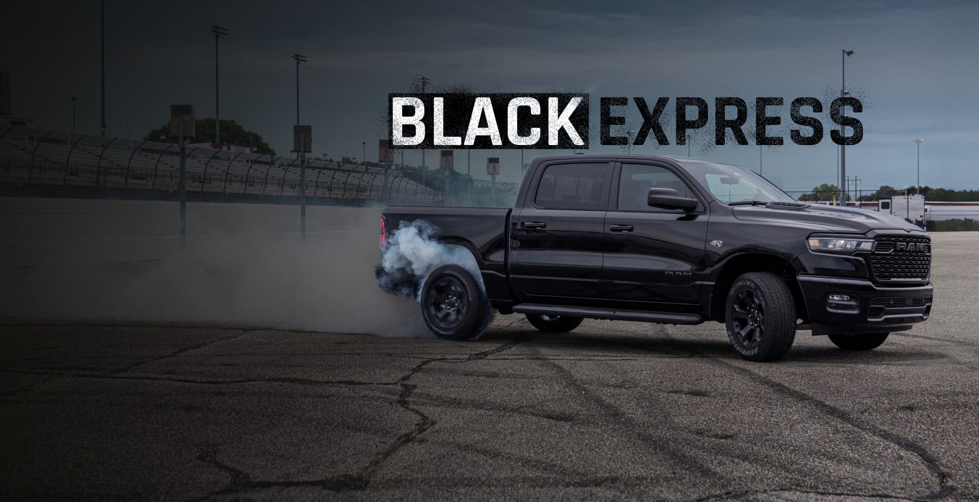 An angled passenger-side profile of a black 2026 Ram 1500 Black Express Crew Cab at a racetrack with smoke billowing from its rear tires. Black Express.