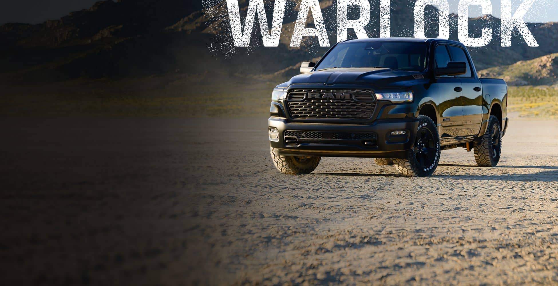 An angled front shot of a black 2026 Ram 1500 Warlock Crew Cab parked on a sandy clearing in the desert. Warlock.