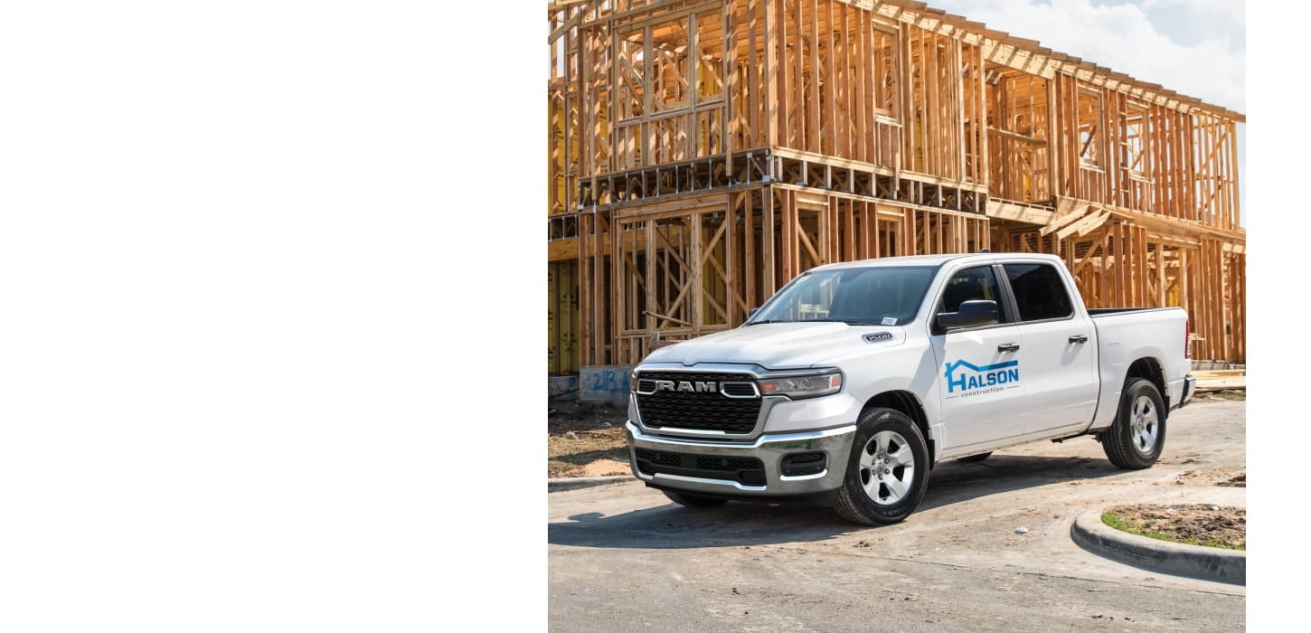 A white 2026 Ram 1500 Tradesman 4x4 Crew Cab with a construction company's signage on its driver's door, parked at a building site.