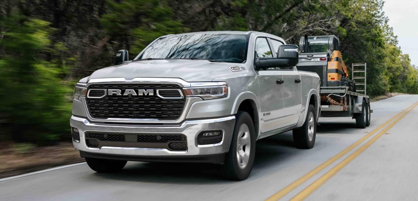 A silver 2026 Ram 1500 Big Horn 4x4 Crew Cab towing motorized equipment as it travels down a highway.