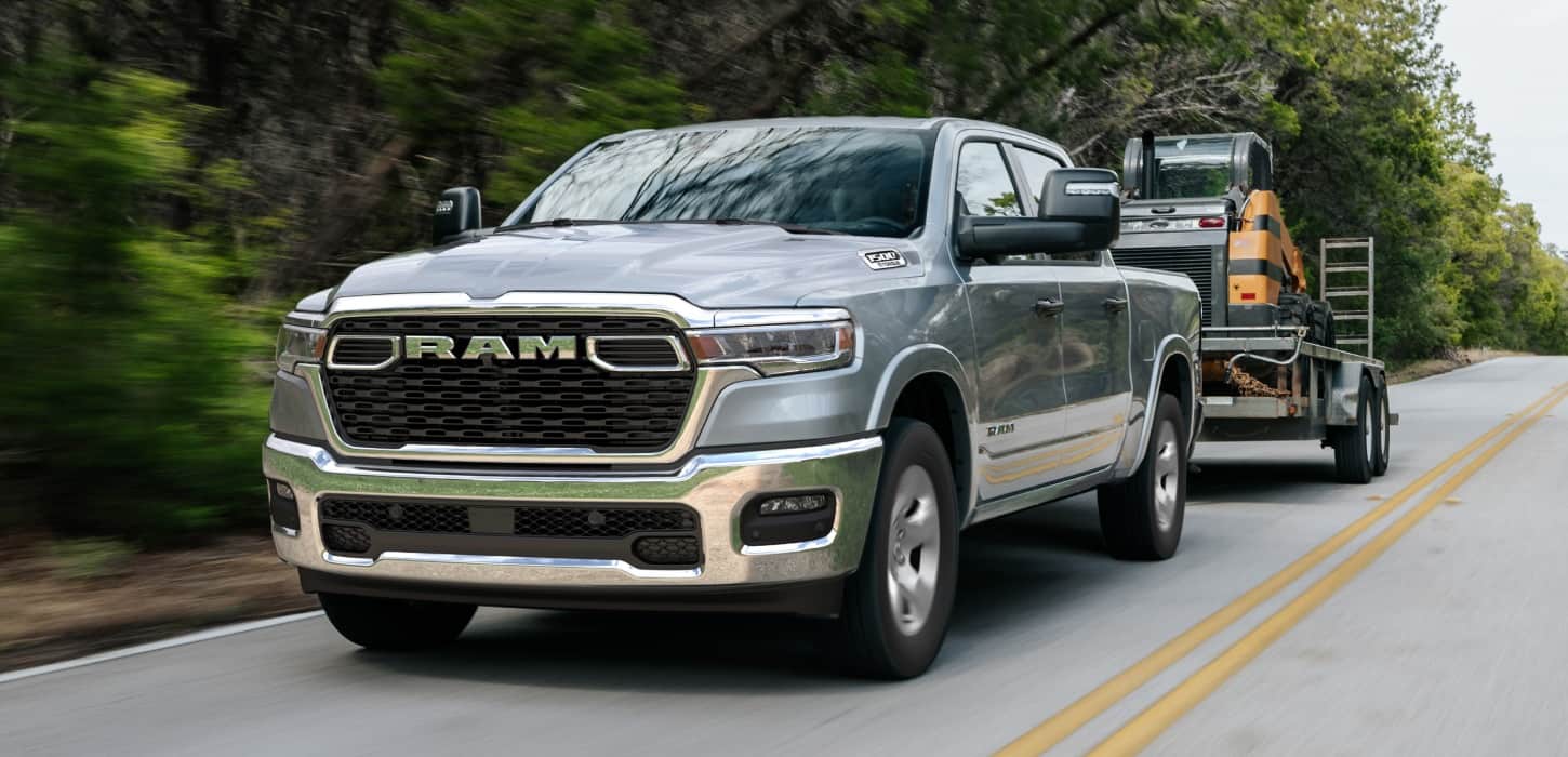 A silver 2026 Ram 1500 Big Horn 4x4 Crew Cab towing motorized equipment as it travels down a highway.