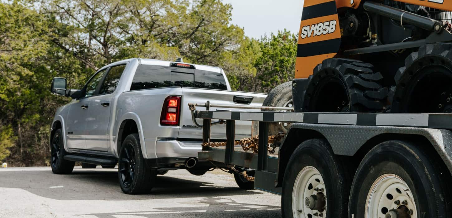 A rear angle of a white 2026 Ram 1500 Laramie 4x4 Crew Cab towing motorized equipment on a utility trailer.