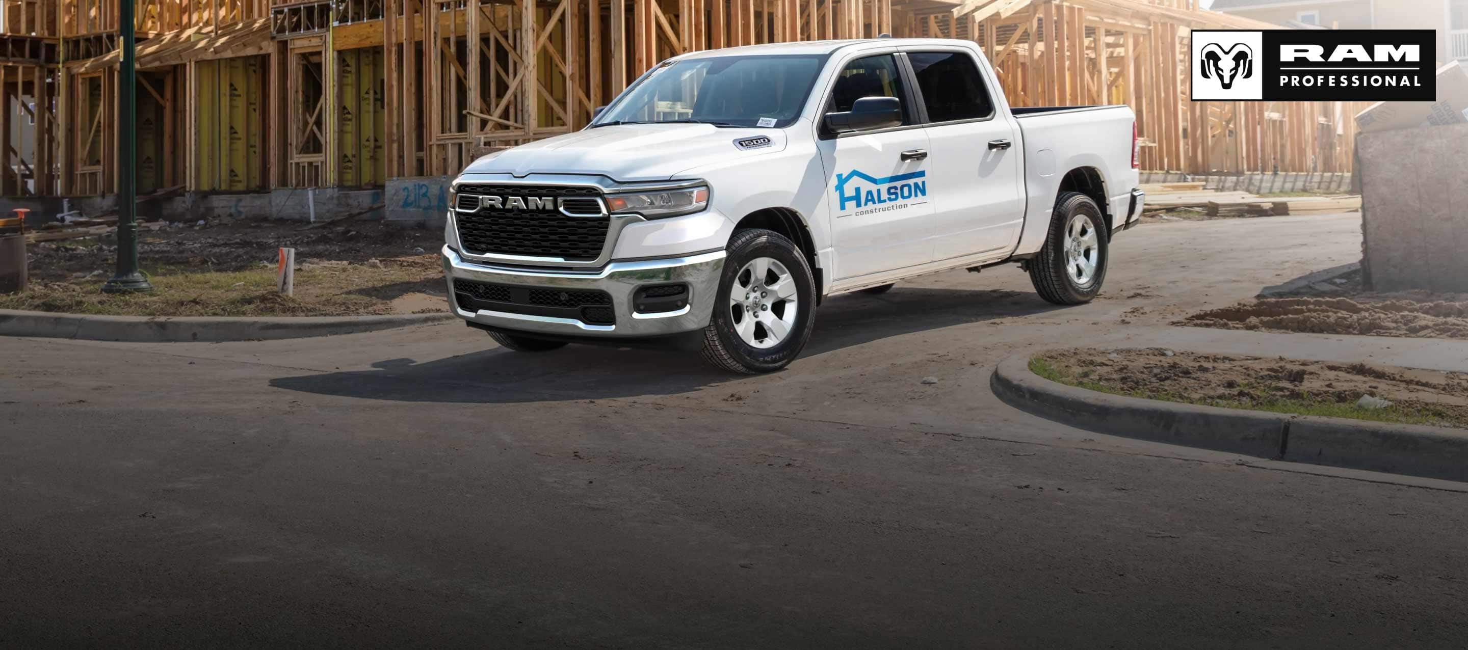 A white 2026 Ram 1500 Tradesman 4x4 Crew Cab, with construction company signage on its driver's door as it pulls away from a commercial building site. Ram Professional.