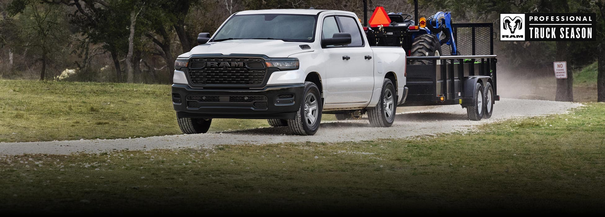 A white 2026 Ram 1500 Tradesman 4x4 Crew Cab towing a tractor on a landscape trailer as it travels down a dirt road. Ram Professional Truck Season.
