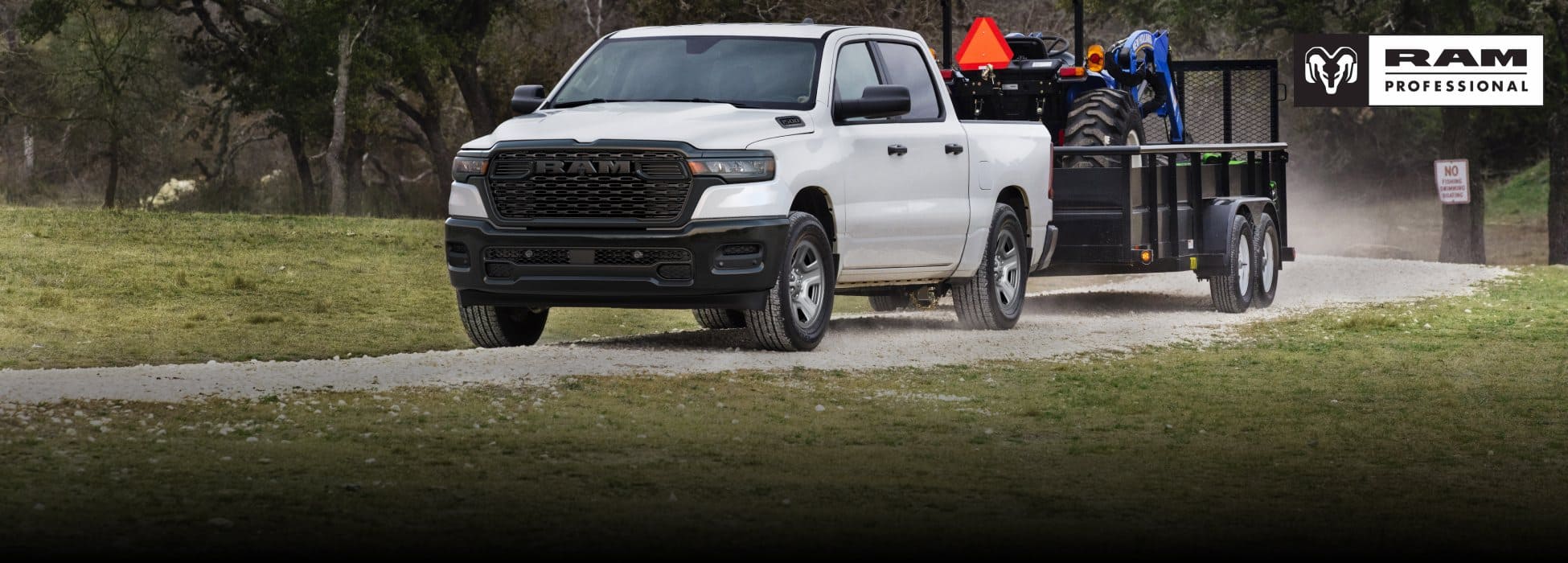 A white 2026 Ram 1500 Tradesman 4x4 Crew Cab towing a tractor on a landscape trailer as it travels down a dirt road. Ram Professional.