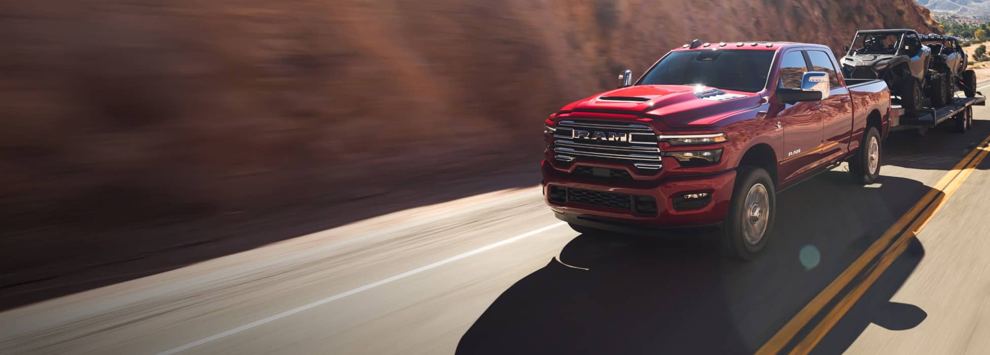 A red 2026 Ram 2500 Laramie 4x4 Crew Cab traveling on a highway in the mountains, while towing two ATVs.