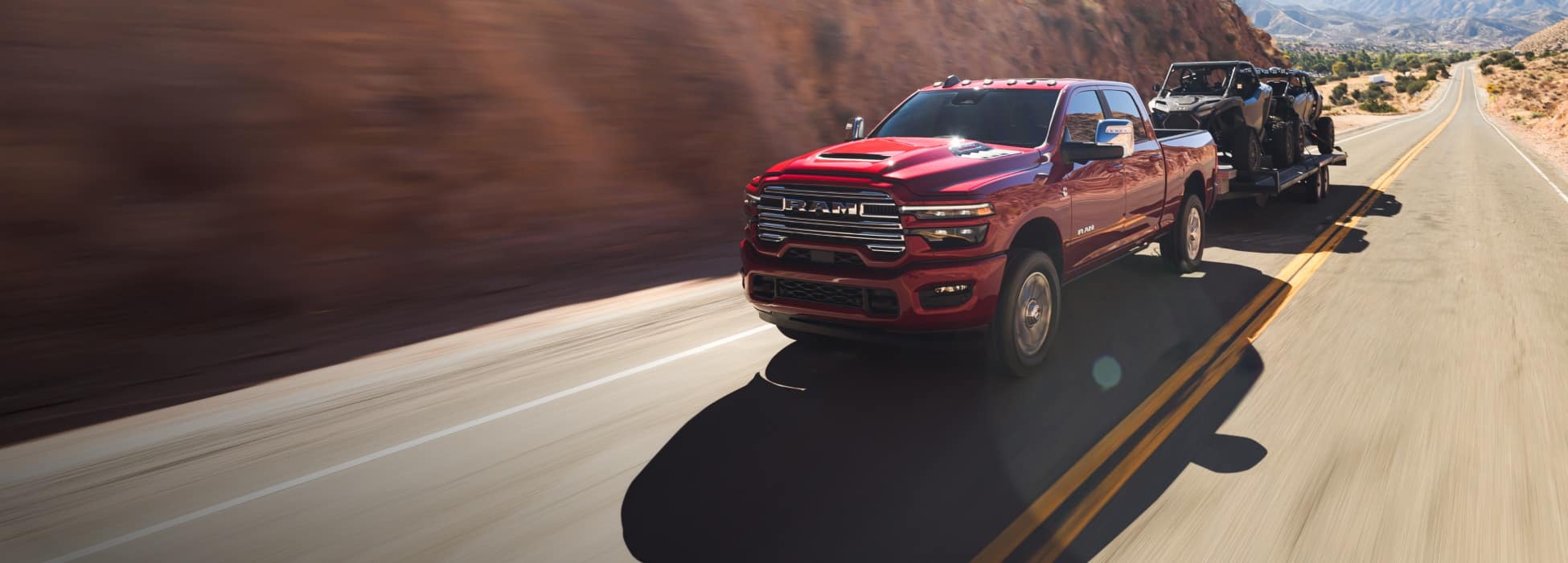A red 2026 Ram 2500 Laramie 4x4 Crew Cab traveling on a highway in the mountains, while towing two ATVs.
