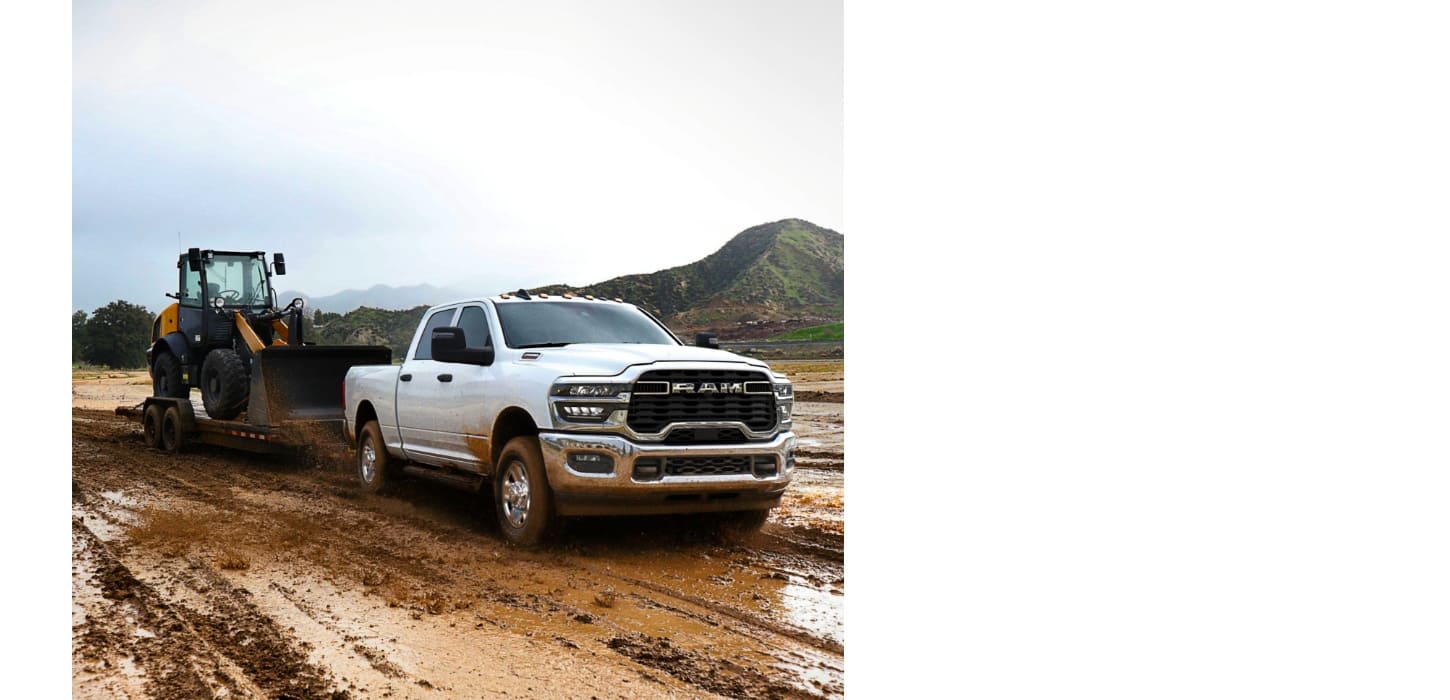 A white 2026 Ram 2500 Tradesman Crew Cab heavy duty truck towing a front end loader on a flatbed trailer through a muddy construction site