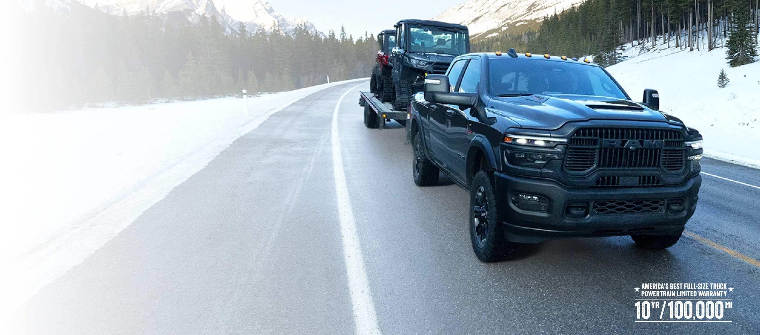 A black 2026 Ram 2500 Rebel 4x4 Crew Cab traveling down a highway in winter, towing two earth moving vehicles. America's Best Full-Size Truck Powertrain Limited Warranty—10-Year/100,000-Mile.