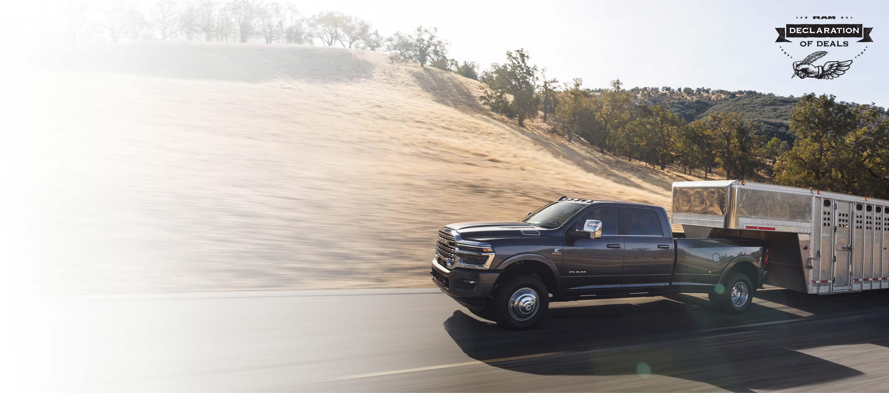 A gray 2026 Ram 3500 Limited Longhorn 4x4 Crew Cab towing a large fifth-wheel livestock trailer as it travels down a highway. Declaration of Deals Sales Event.