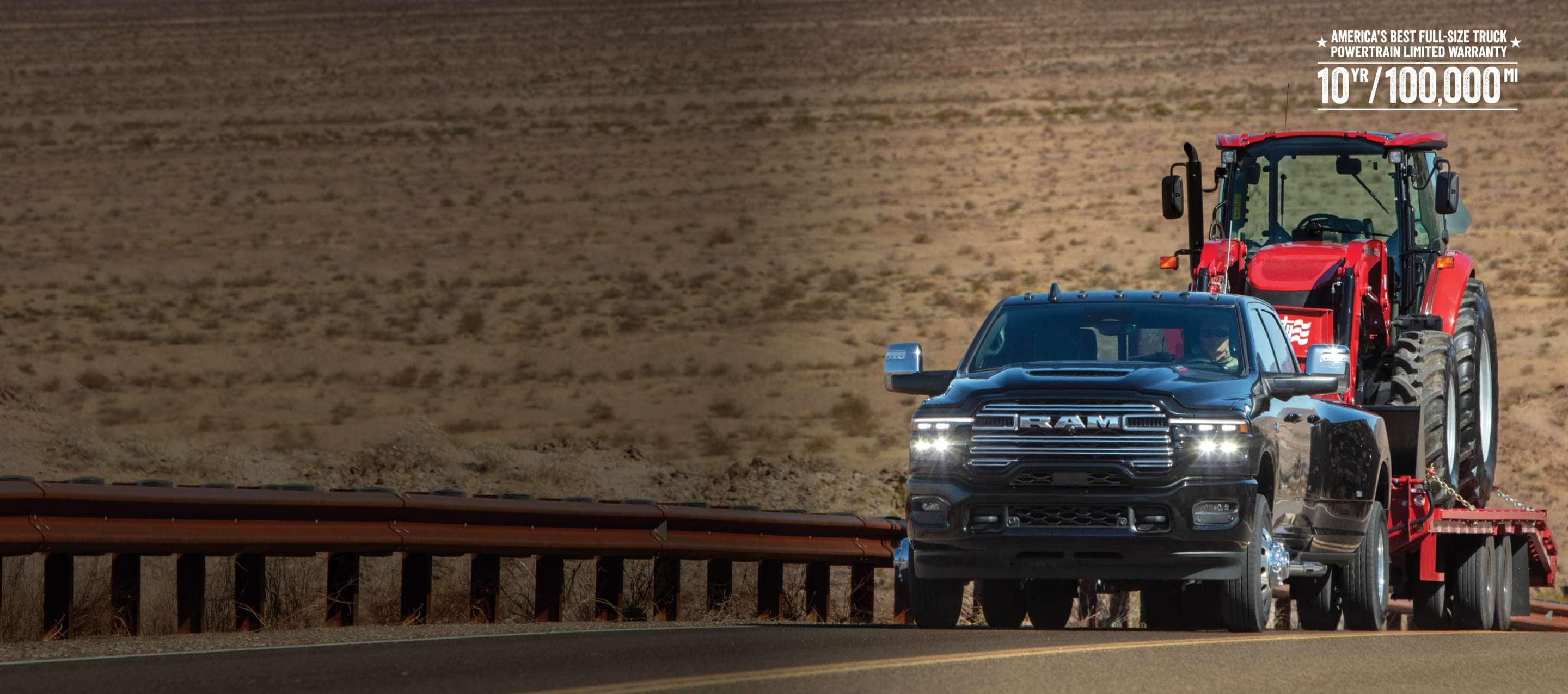 A black 2026 Ram 3500 Laramie 4x4 Crew Cab towing a construction vehicle on a long flatbed trailer as it travels up a desert highway. America's Best Full-Size Truck Powertrain Limited Warranty—10-Year/100,000-Mile.