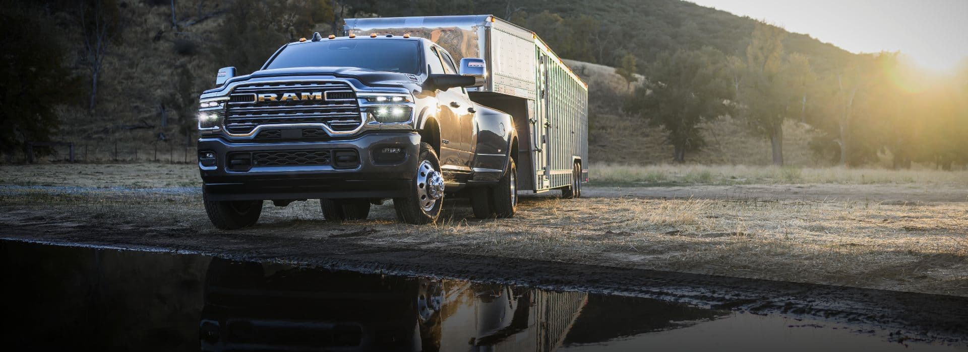 A black 2026 Ram 3500 Limited Longhorn 4x4 Crew Cab towing a large fifth-wheel livestock trailer off-road, beside a waterfront.