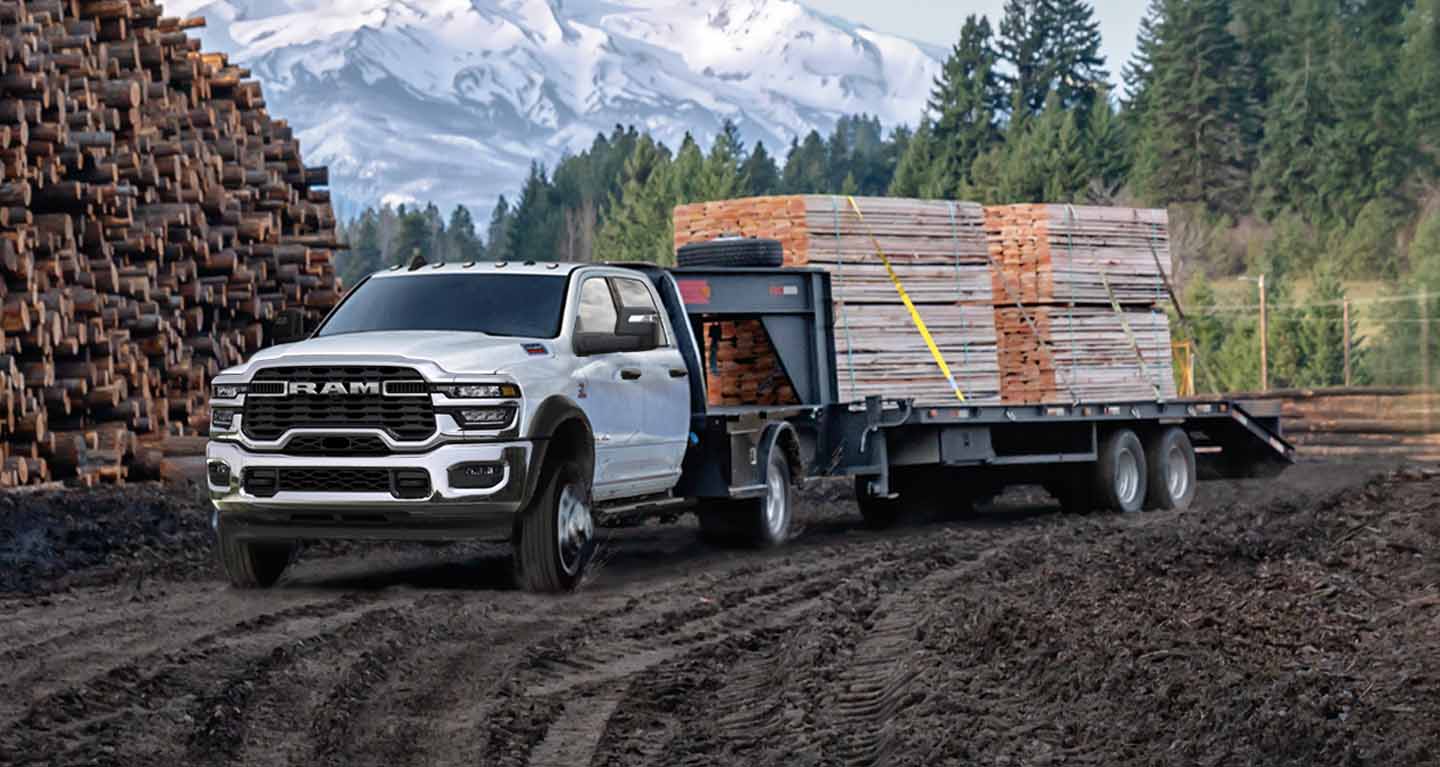 Display A white 2026 Ram 5500 Tradesman Chassis Cab Crew Cab with a gooseneck trailer filled with lumber, parked beside a forest and huge mound of cut logs.