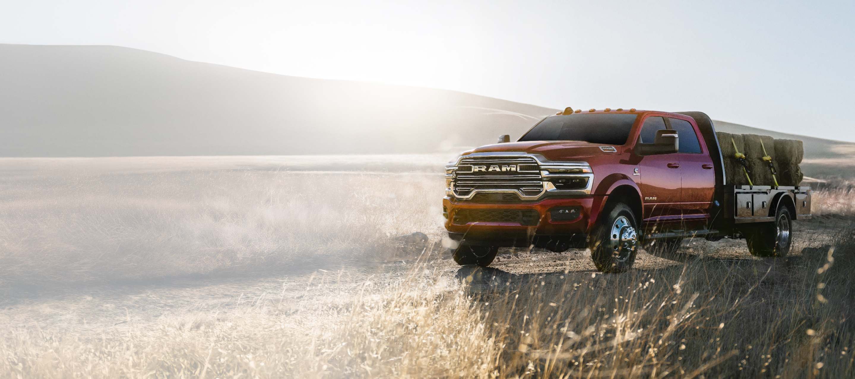 A red 2026 Ram 5500 Big Horn Chassis Cab 4x4 Crew Cab, with the Laramie Trim Group, traveling on a dirt road in the desert, towing several large bales of hay on a platform upfit. Ram Professional