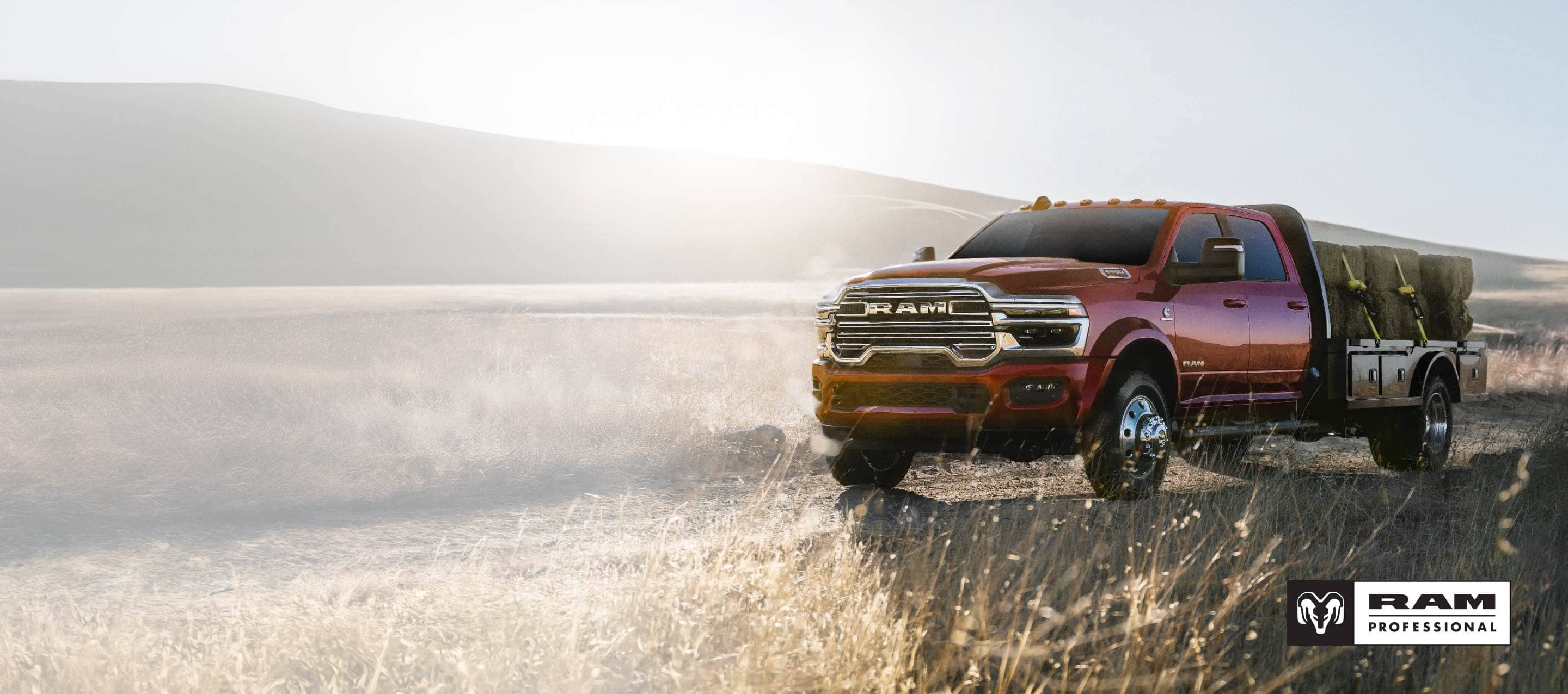 A red 2026 Ram 5500 Big Horn Chassis Cab 4x4 Crew Cab, with the Laramie Trim Group, traveling on a dirt road in the desert, towing several large bales of hay on a platform upfit. Ram Professional