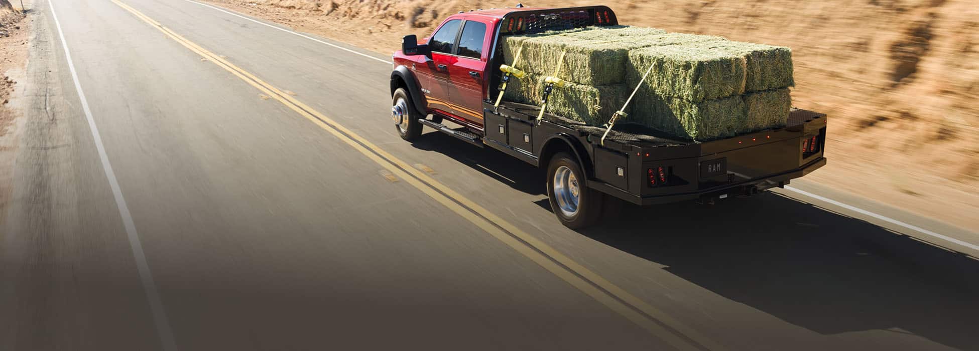 A rear angle of a red 2026 Ram 4500 Big Horn Chassis Cab Crew Cab traveling down a highway, carrying several large bales of hay on a long platform upfit.