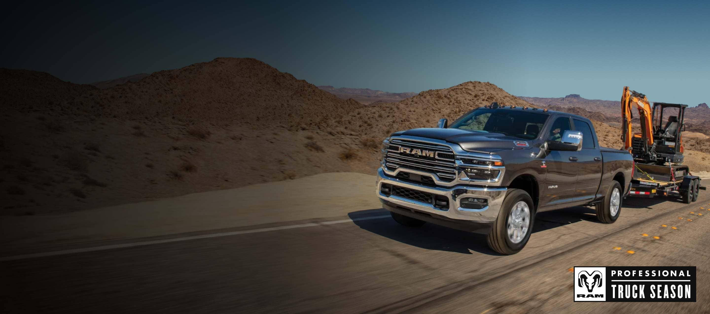 A black 2026 Ram 2500 Laramie 4x4 Crew Cab traveling down a highway, towing an excavator on a flat bed trailer. Ram Professional Truck Season.