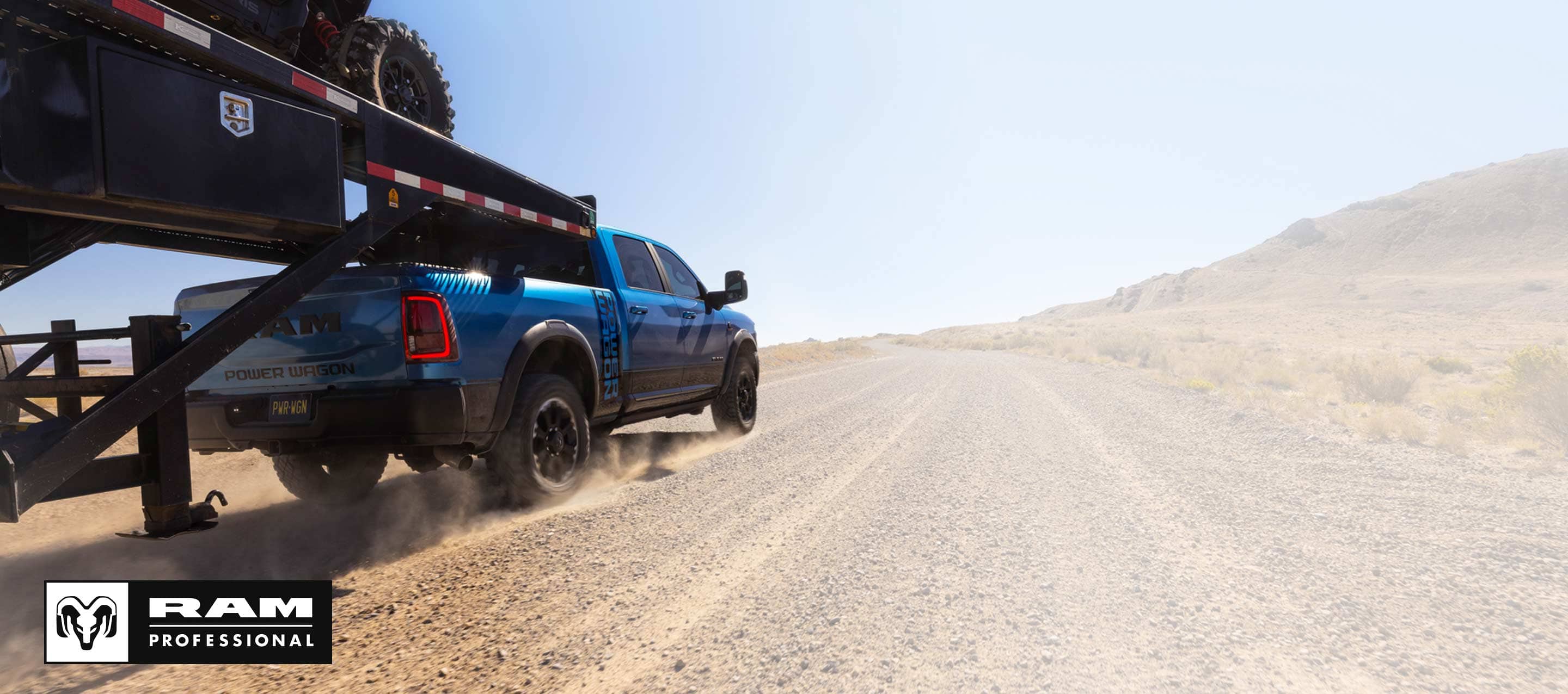 A blue 2026 Ram 2500 Heavy Duty Power Wagon 4x4 Crew Cab towing an ATV on a fifth-wheel flatbed trailer as it travels down a dirt road. Ram Professional.