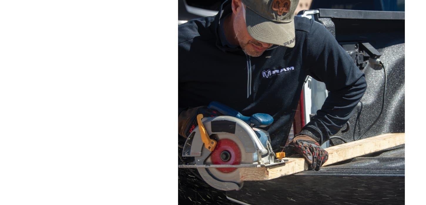 An open pickup bed on a 2026 Ram 2500 heavy duty truck with a man cutting a piece of plywood with a circular power saw plugged into the pickup's onboard power.