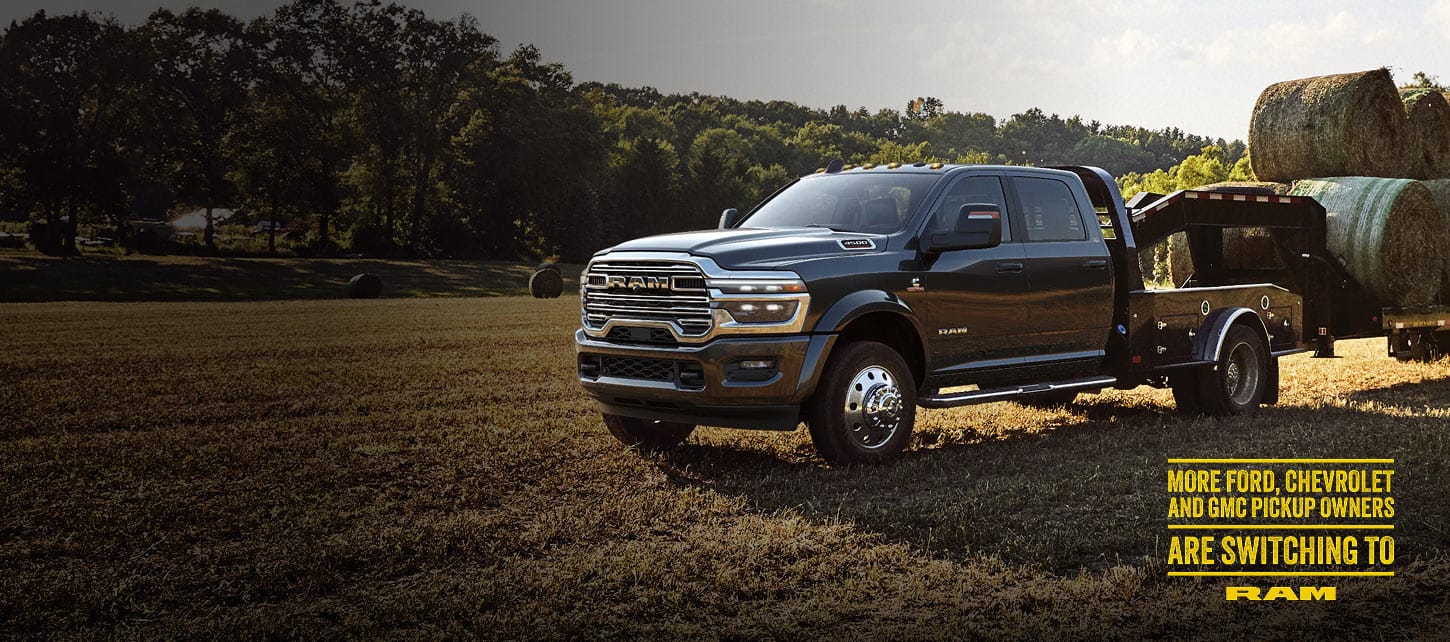 A black 2026 Ram 4500 Big Horn Chassis Cab Crew Cab with the Laramie Trim Group and a gooseneck trailer loaded with several large bales of hay, parked in an open field. 