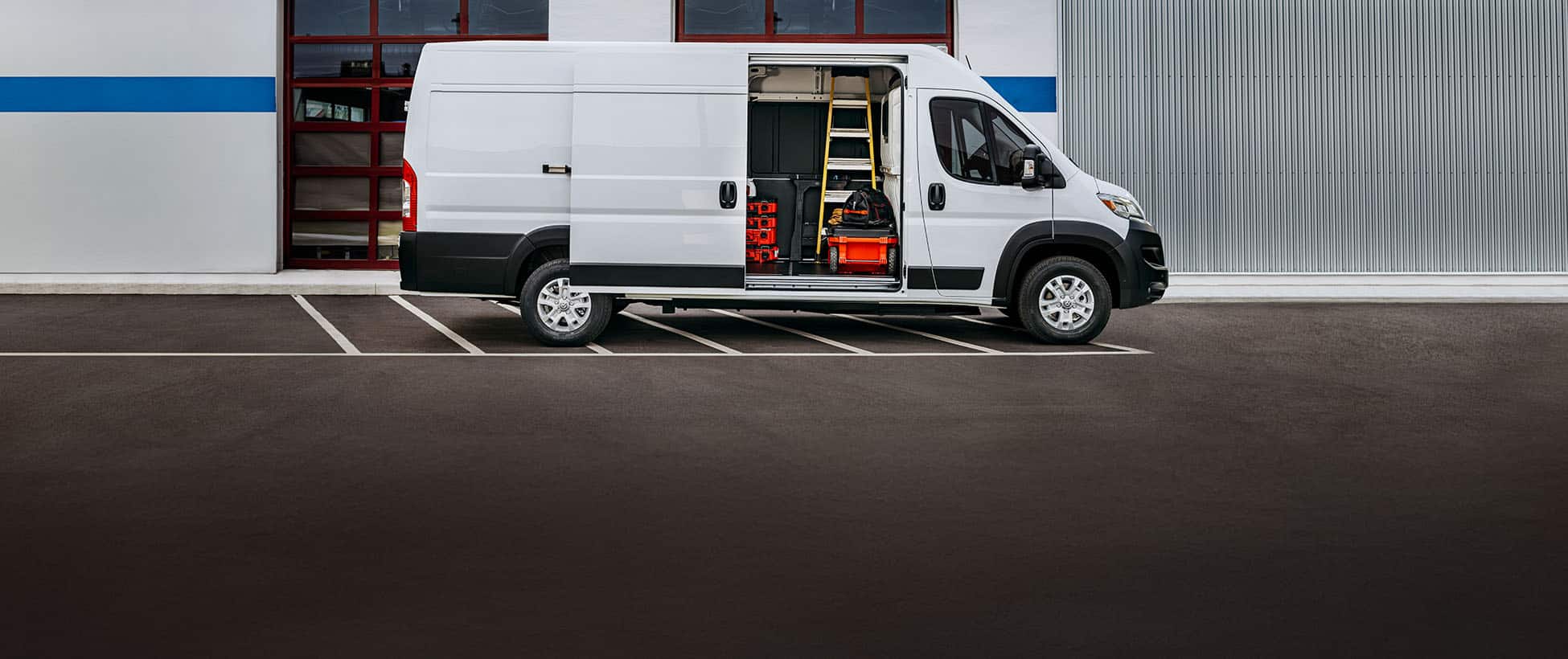 A passenger-side profile of a white 2026 Ram ProMaster EV 3500 SLT High Roof Cargo Van parked beside a commercial garage with its sliding side door open, revealing tools, equipment and a ladder inside.
