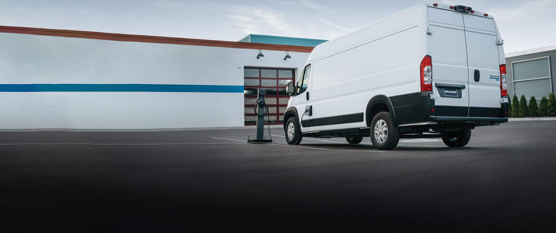 A white 2026 Ram ProMaster EV 3500 SLT High Roof Cargo Van parked beside a public charging station with its charging cord plugged into the van's charging port.