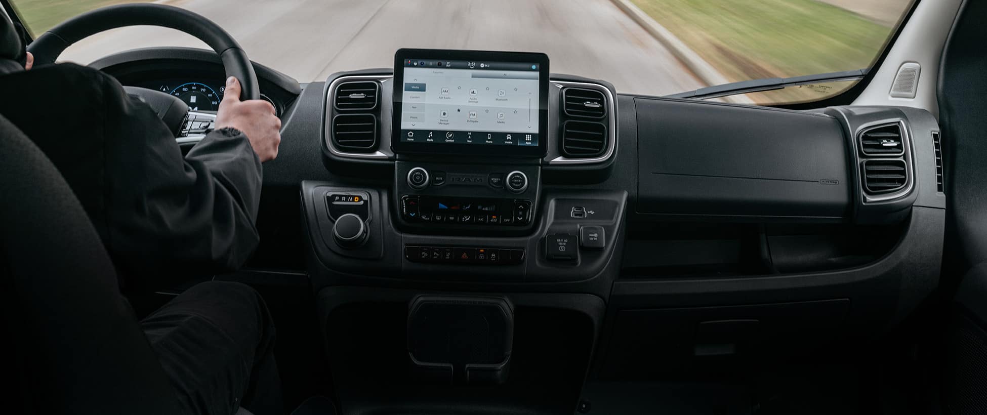 The front seat area in a 2026 Ram ProMaster EV 3500 Cargo Van with a driver traveling through a residential neighborhood and the Uconnect touchscreen displaying media selections.