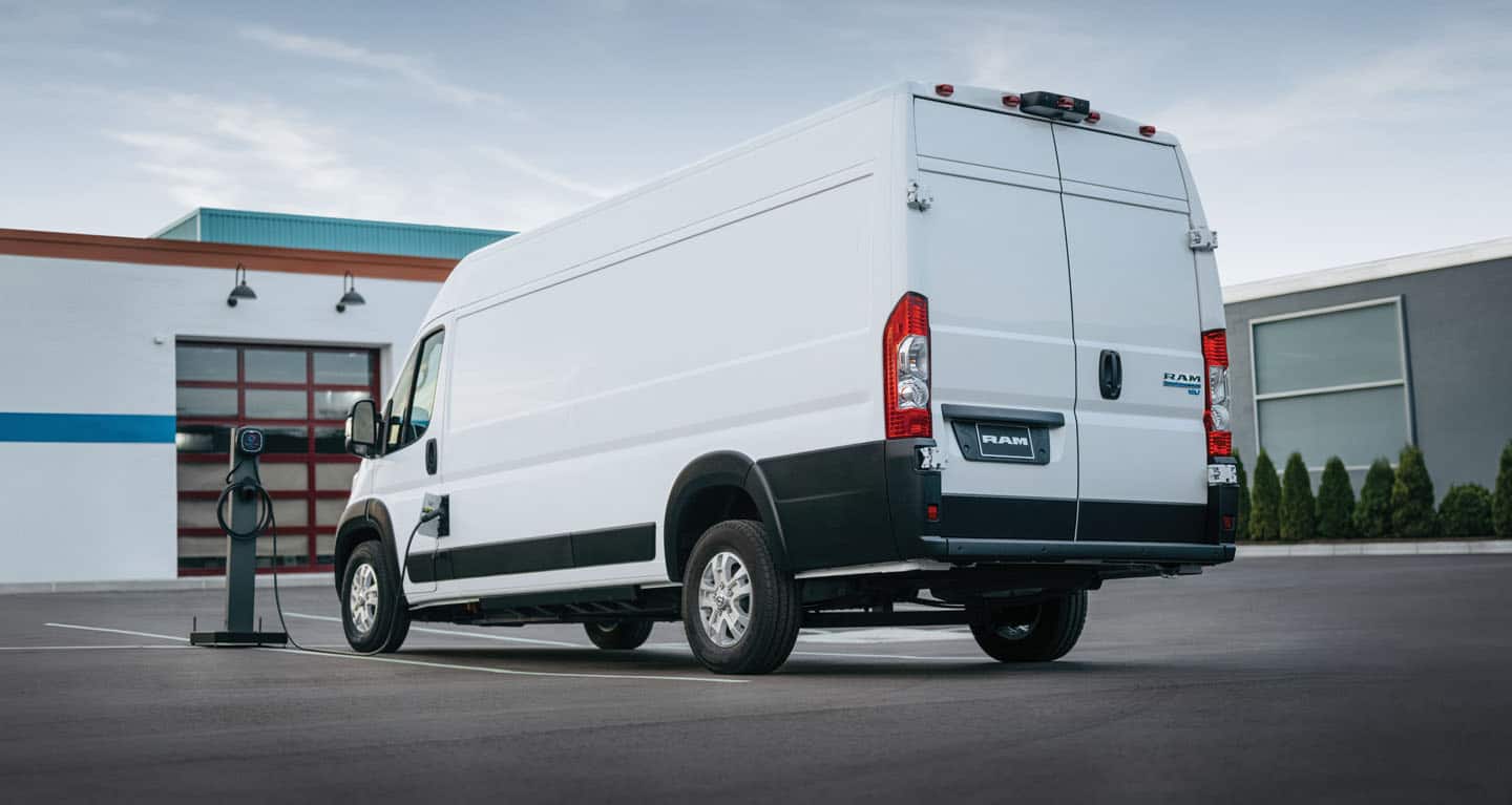 Display A white 2026 Ram ProMaster EV 3500 SLT High Roof Cargo Van parked beside a public charging station with its charging cord plugged into the van's charging port.