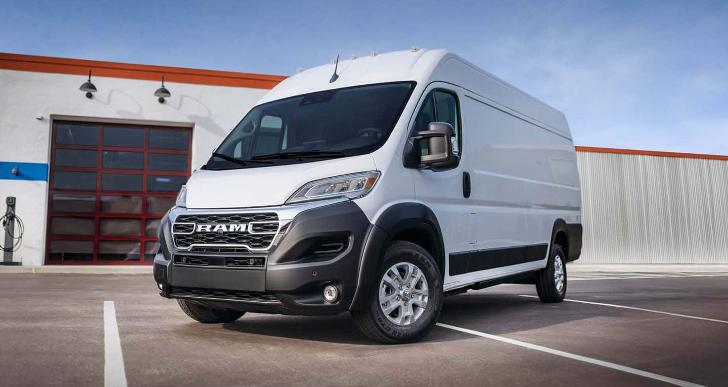 Display A driver-side front angle of a white 2026 Ram ProMaster EV 3500 SLT High Roof Cargo Van parked near a commercial garage.