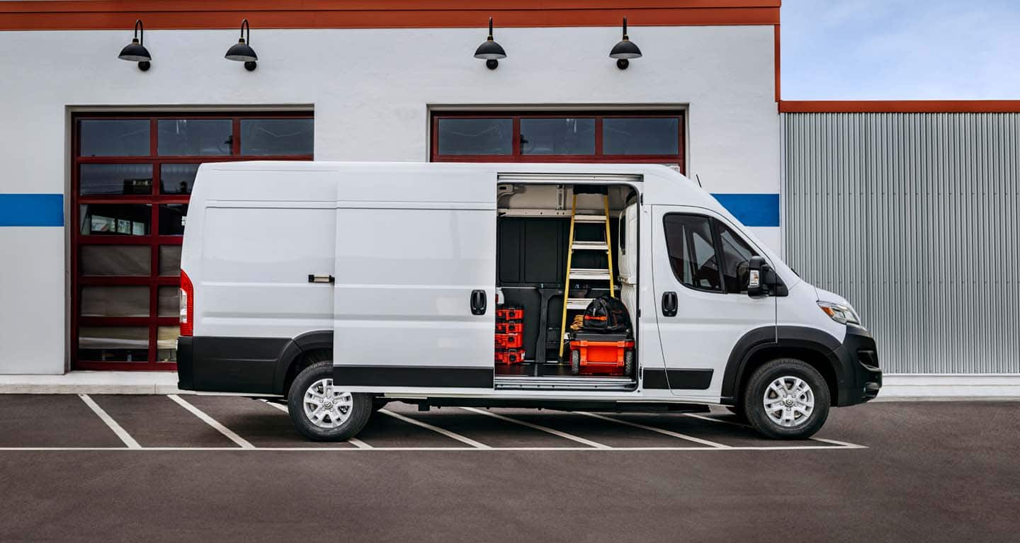 Display A passenger-side profile of a white 2026 Ram ProMaster EV 3500 SLT High Roof Cargo Van parked beside a commercial garage with its sliding side door open, revealing tools, equipment and a ladder inside.