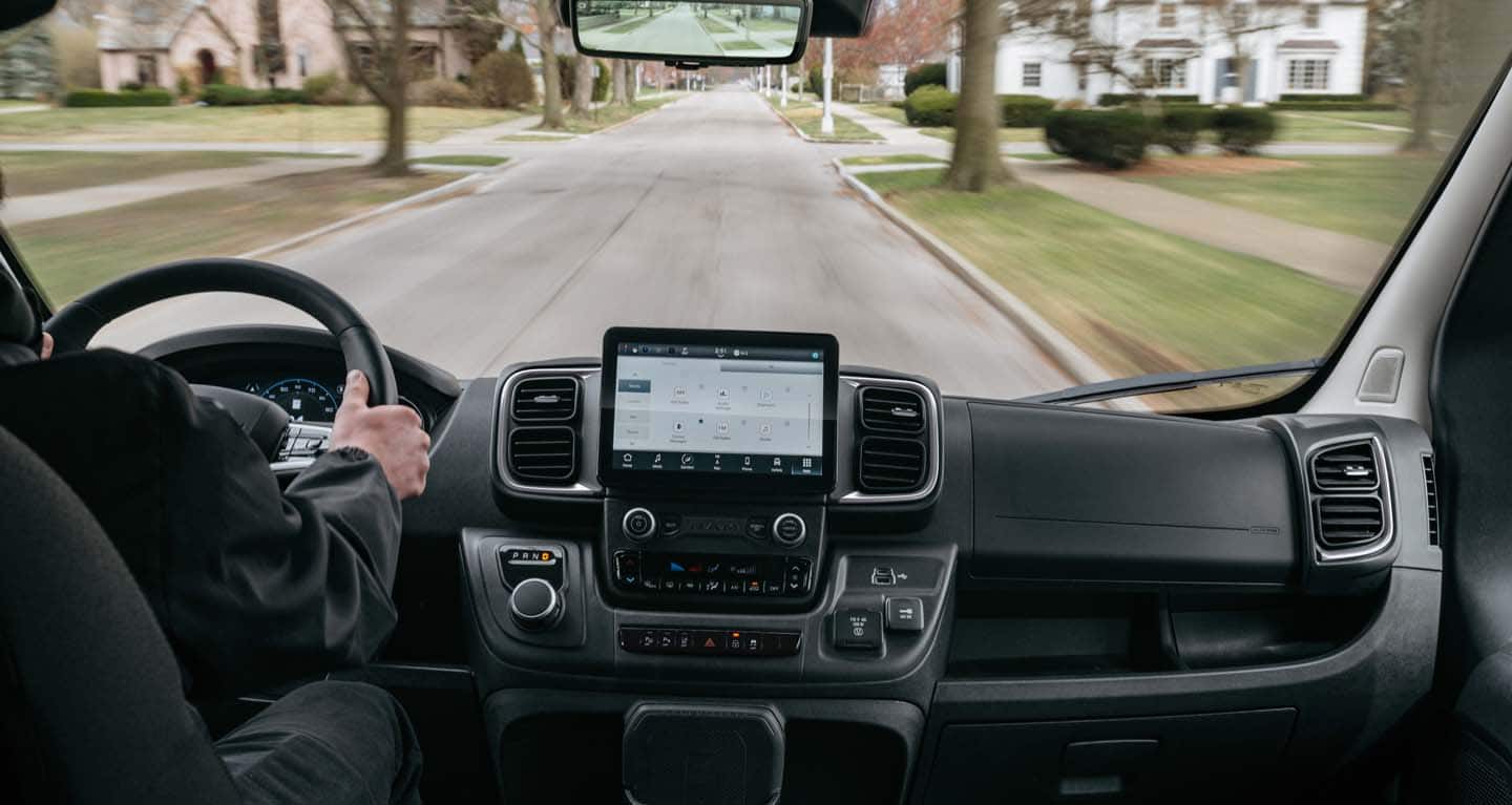Display The front seat area in a 2026 Ram ProMaster EV 3500 Cargo Van with a driver traveling through a residential neighborhood and the Uconnect touchscreen displaying media selections.