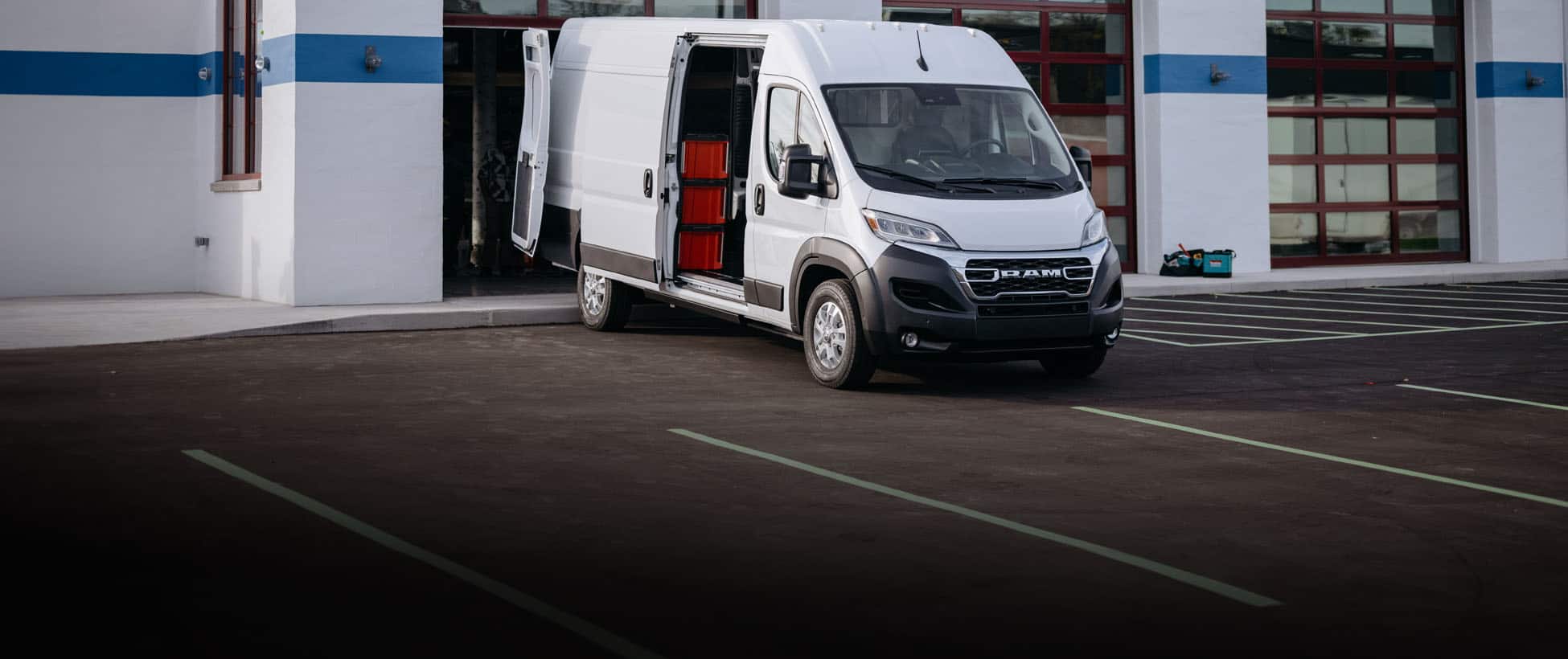A passenger-side front angle of a white 2026 Ram ProMaster EV 3500 SLT High Roof Cargo Van with its side and rear doors open, parked near a commercial building's open garage door.