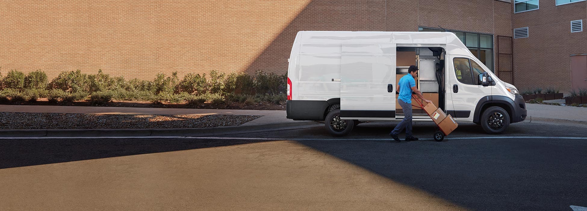 A man pushing packages on a dolly near the open sliding side door on a white 2026 Ram ProMaster 3500 SLT Plus Cargo Van Super High Roof with Black Appearance Package.