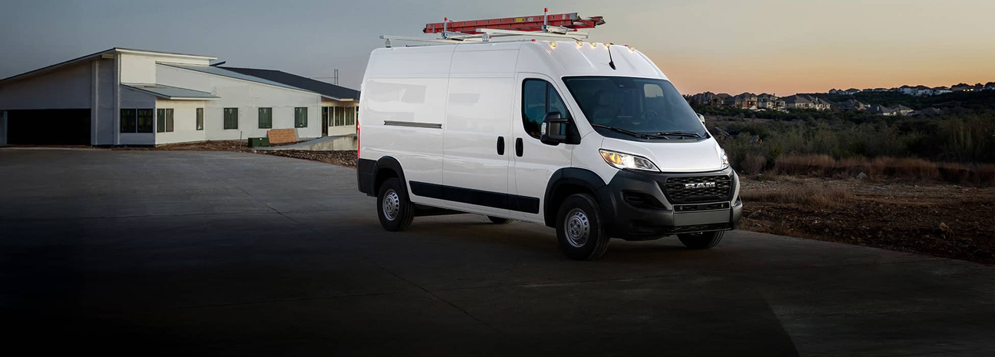 A passenger-side front angle of a white 2026 Ram ProMaster Tradesman Cargo Van High Roof with a ladder on its roof rack leaving a construction site.