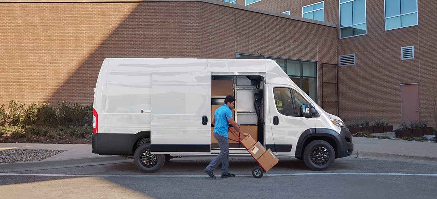 Display A man pushing packages on a dolly near the open sliding side door on a white 2026 Ram ProMaster 3500 SLT Plus Cargo Van Super High Roof with Black Appearance Package.