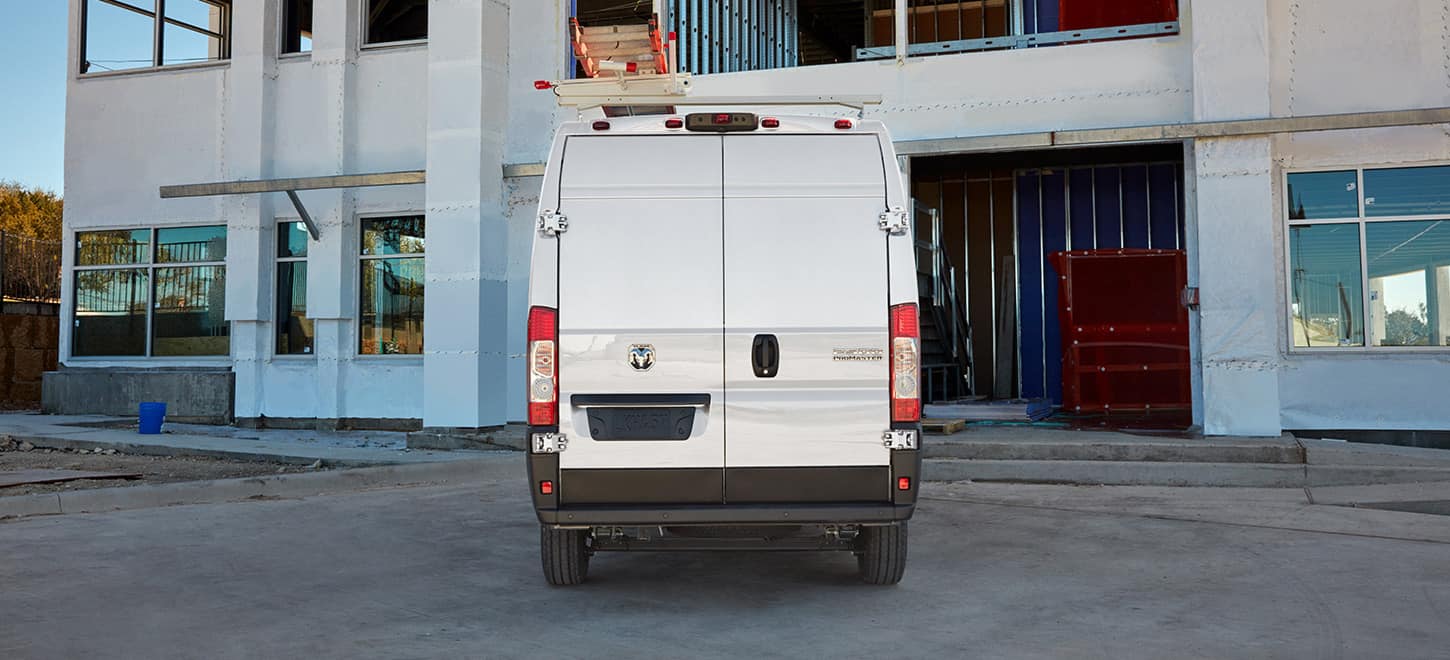 Display The rear of a white 2026 Ram ProMaster 2500 Cargo Van High Roof, with a ladder rack, pulling into the garage of a commercial building.