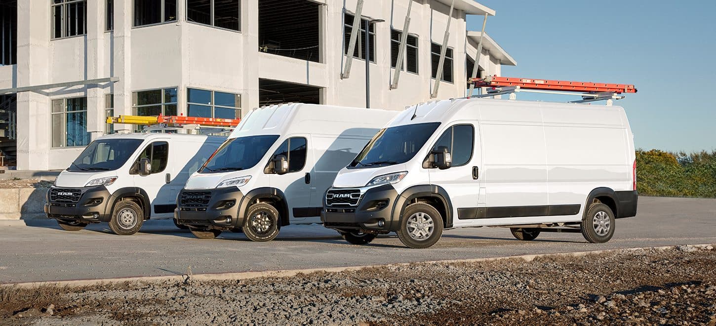 Display A lineup of three white 2026 Ram ProMaster vans parked at a construction site: from left to right, a Ram ProMaster 1500 Tradesman Cargo Van Standard Roof, a Ram ProMaster 3500 SLT Plus Cargo Van Super High Roof with the Black Appearance Package, and a Ram ProMaster 2500 SLT Cargo Van High Roof.