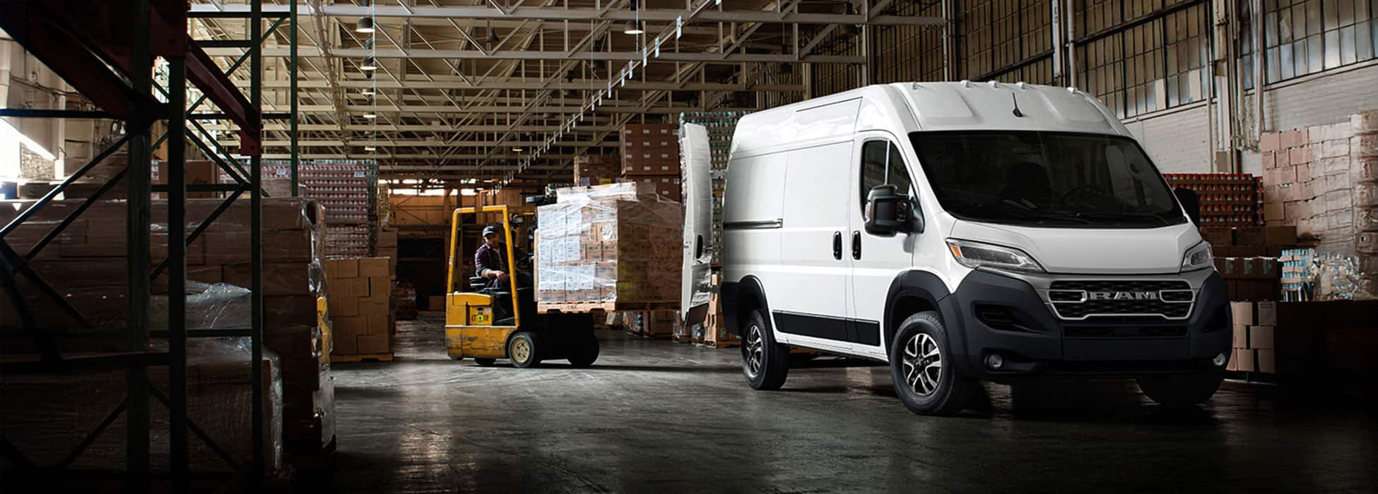 A passenger-side front angle of a white 2026 Ram ProMaster 2500 SLT Cargo Van High Roof, parked in a warehouse with its rear doors fully open and a forklift with a pile of boxes being driven toward the back of the van.
