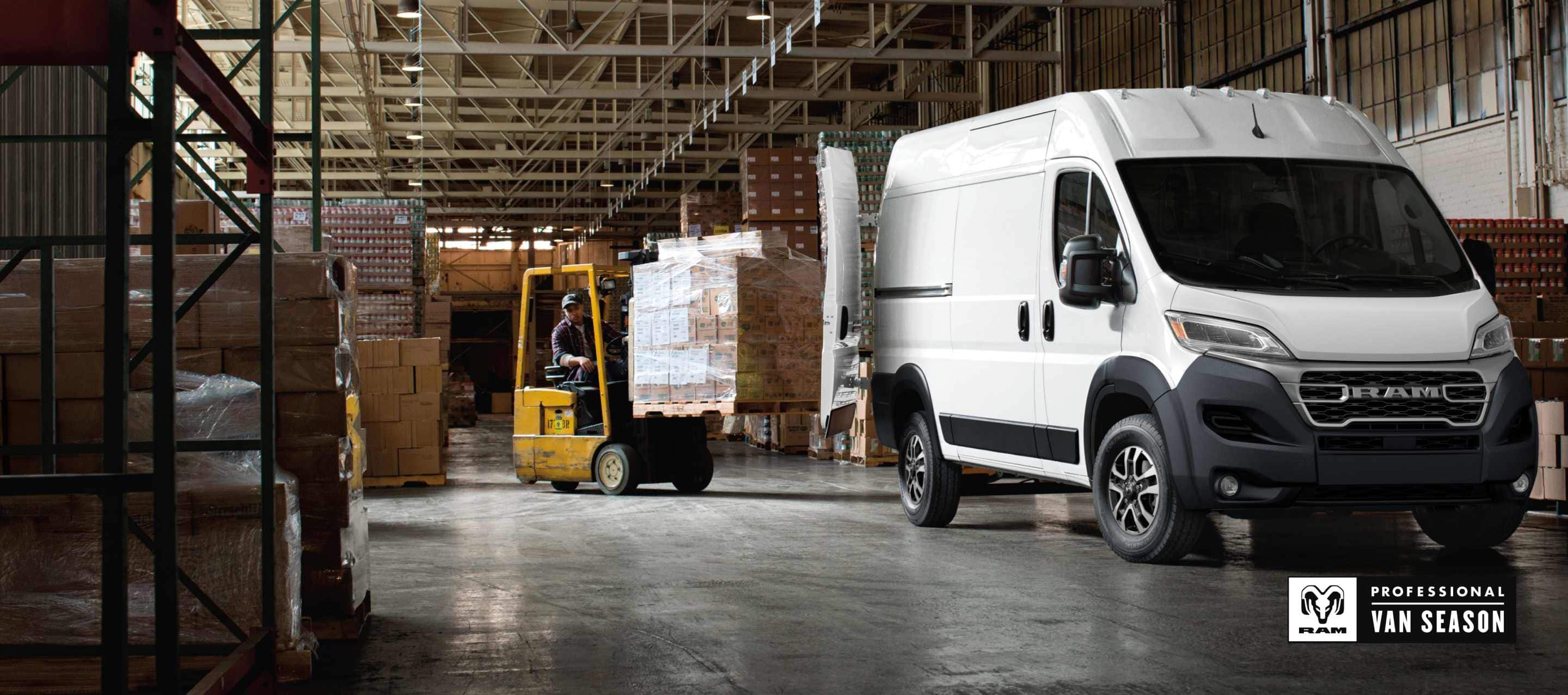 A passenger-side front angle of a white 2026 Ram ProMaster 2500 SLT Cargo Van High Roof, parked in a warehouse with its rear doors fully open and a forklift with a pile of boxes being driven toward the back of the van. Ram Professional Van Season.