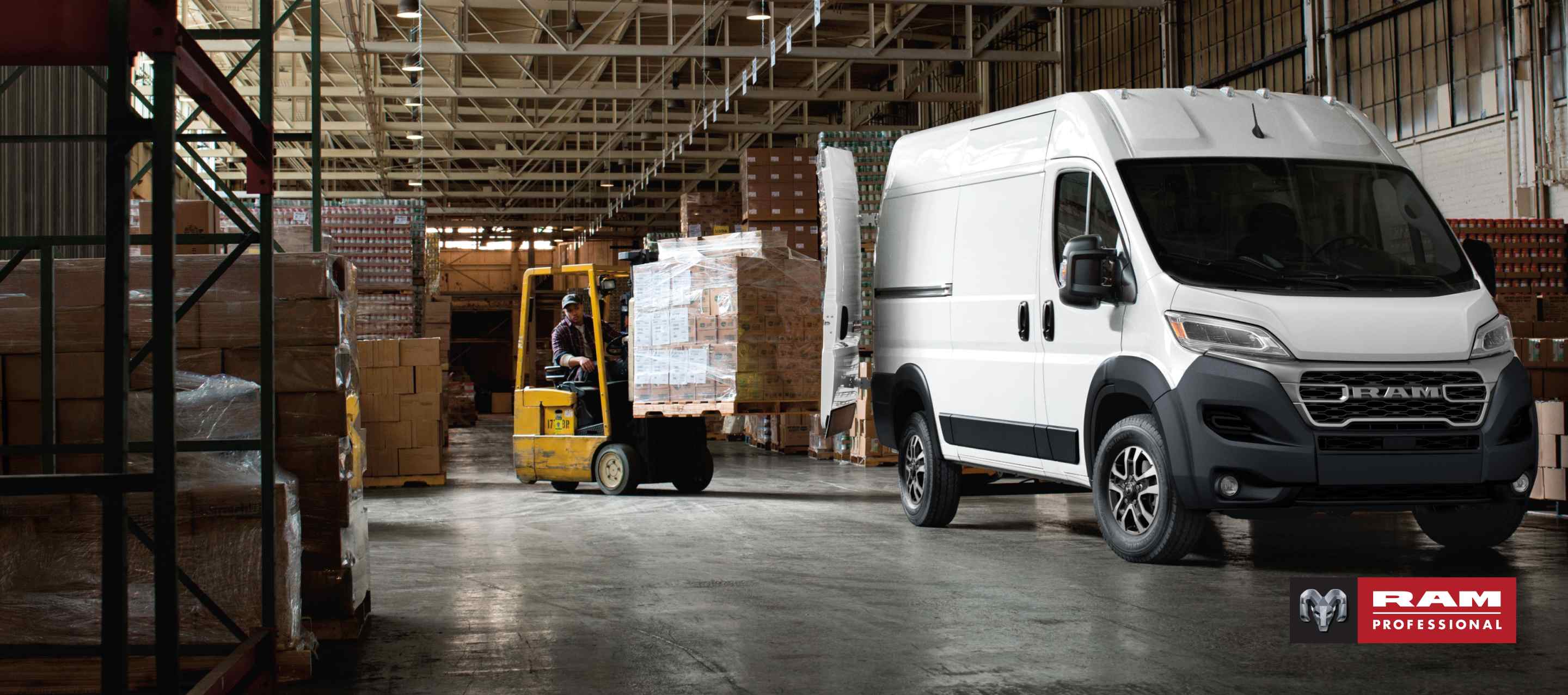 A passenger-side front angle of a white 2026 Ram ProMaster 2500 SLT Cargo Van High Roof, parked in a warehouse with its rear doors fully open and a forklift with a pile of boxes being driven toward the back of the van.