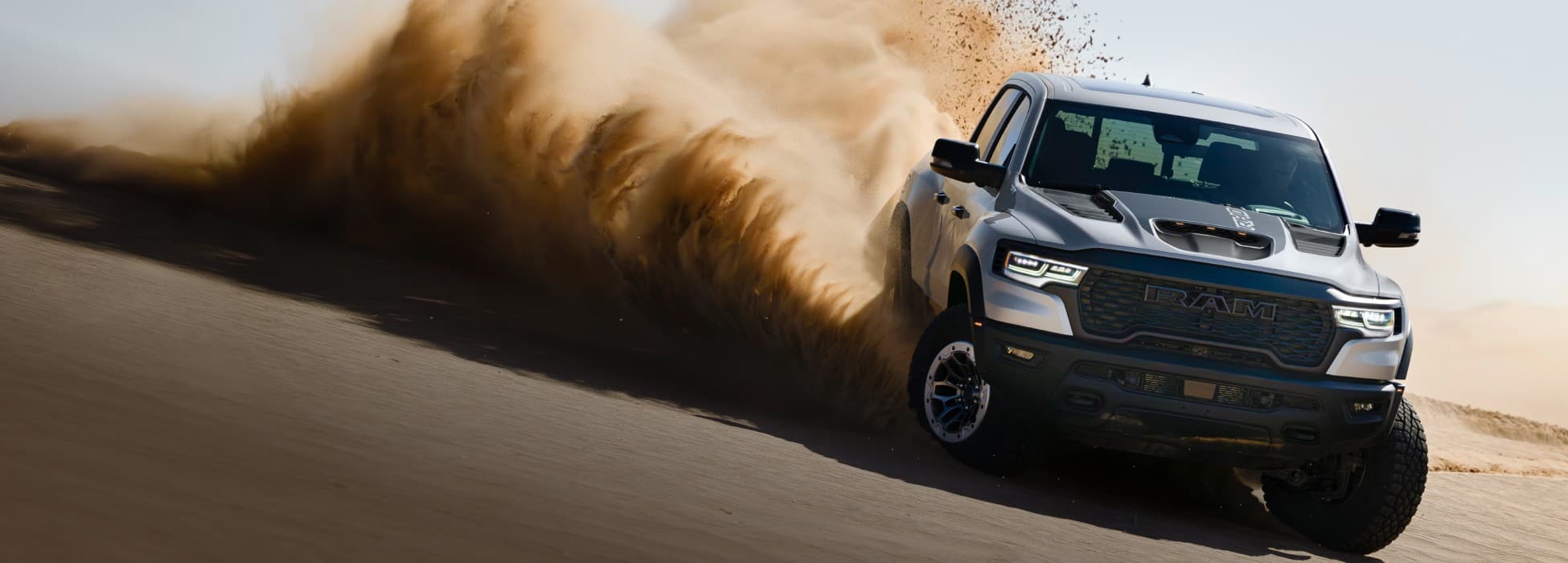 A head-on angle of a silver 2026 Ram 1500 RHO 4x4 Crew Cab traveling off-road on a hill in the desert, with dust and sand billowing from its tires.