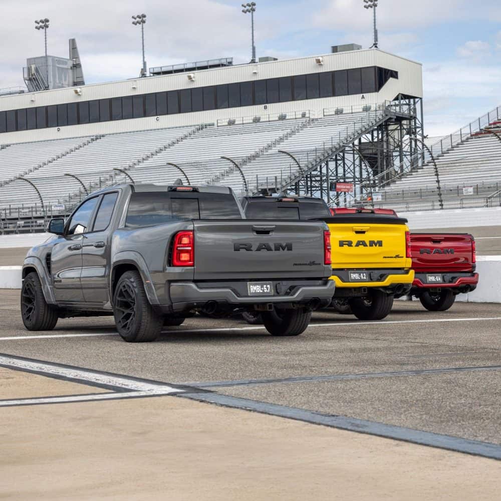 Three 2027 Ram 1500 Rumble Bee trucks in gray, yellow and red at a racetrack, rear view