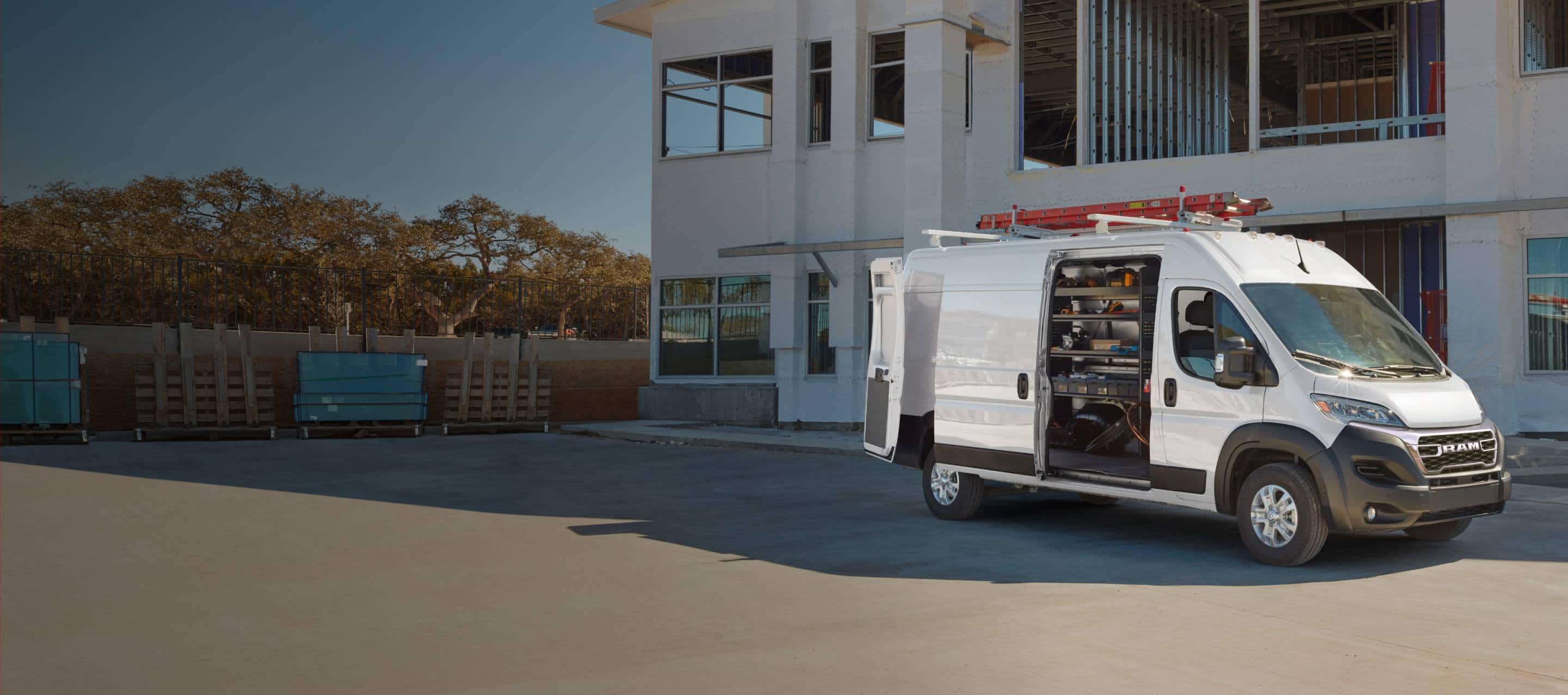 A white 2027 Ram ProMaster 3500 SLT High Roof Cargo Van, parked outside a commercial building with a red ladder on its roof rack and its passenger-side sliding door open, revealing custom shelving full of equipment. Crafted For Professionals. Ram Professional.