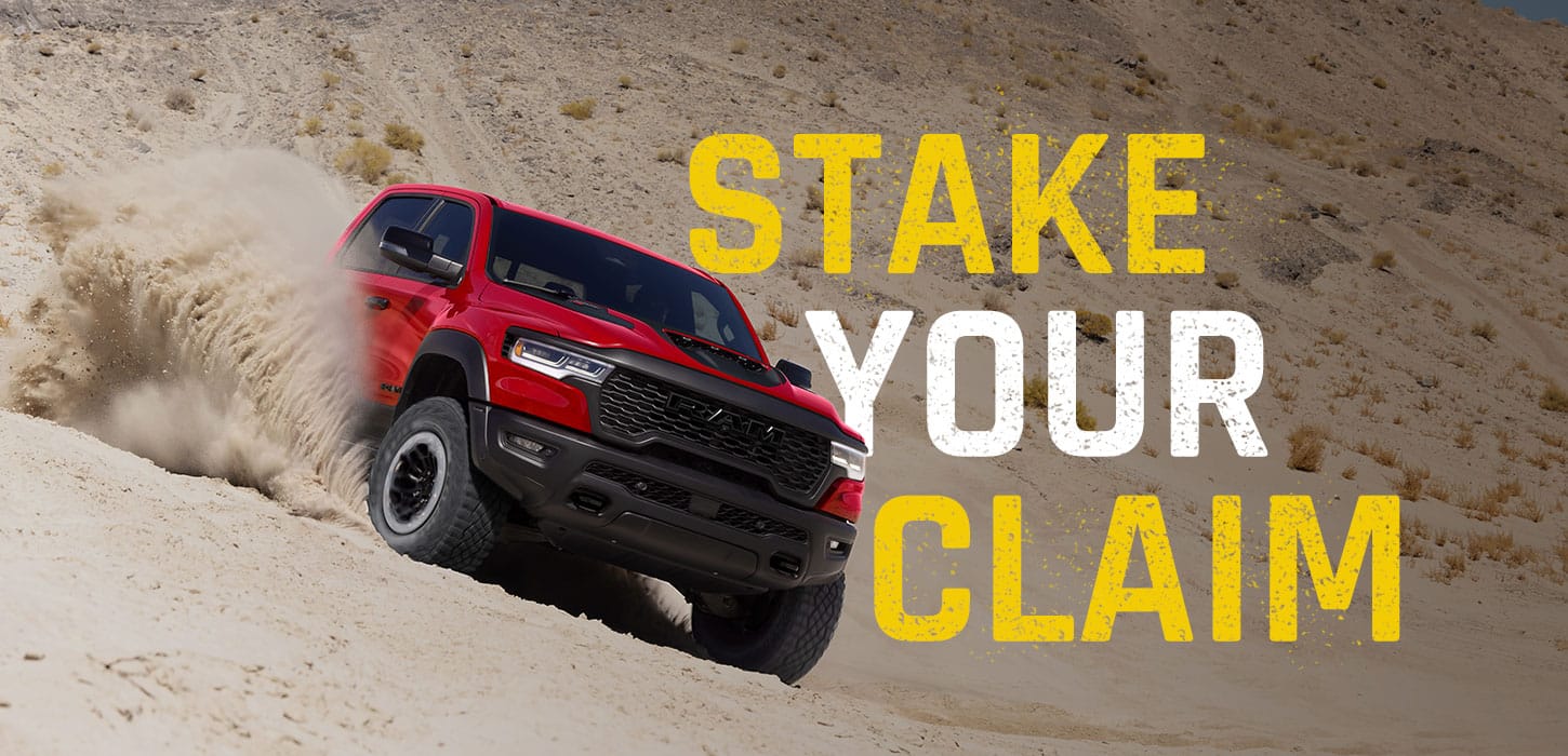 A red 2027 Ram 1500 RHO 4x4 Crew Cab shot descending a dune on an angle, kicking up sand as it goes. Stake Your Claim.