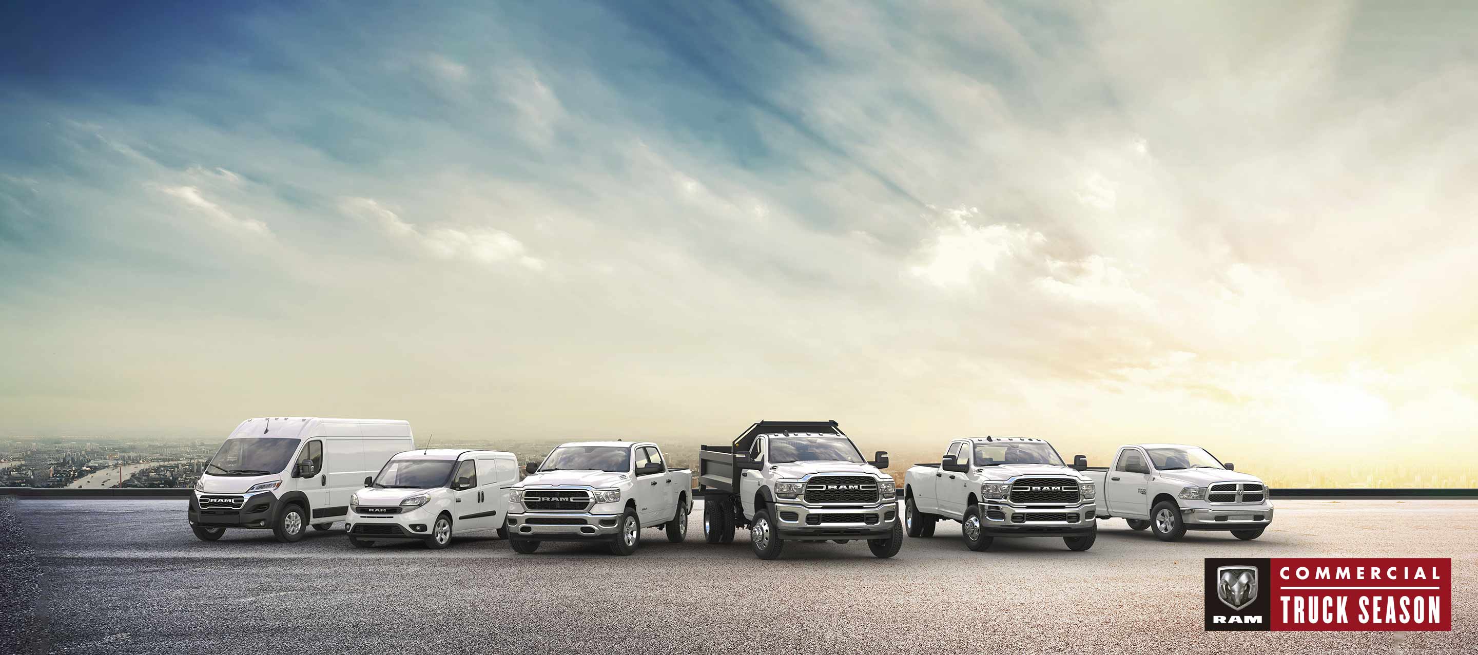 A lineup consisting of a 2023 Ram ProMaster Cargo Van, a 2022 Ram ProMaster City Passenger Van and four 2022 Ram trucks, all parked on a rooftop parking lot with a skyline of highrise buildings in the background. Ram Commercial.