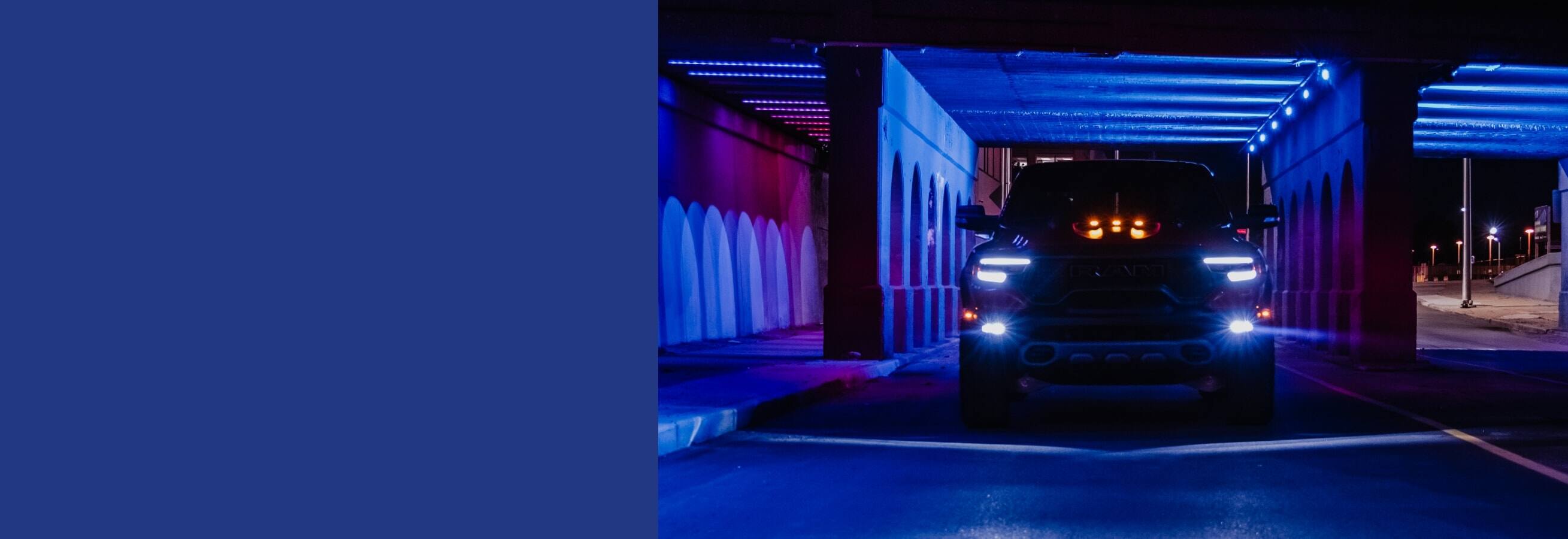 A silhouetted head-on view of a 2022 Ram 1500 TRX with its headlamps on, exiting a tunnel flooded with blue light.